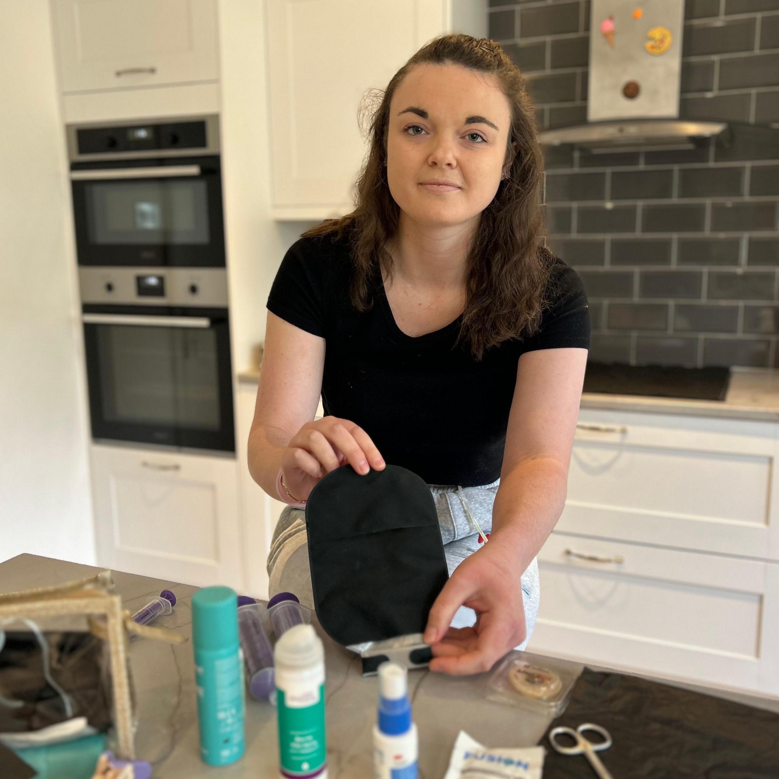 A young woman is sitting on a stool in a modern-looking kitchen holding a black stoma bag. She has dark brown hair and is wearing a black t-shirt and grey jogging bottoms.