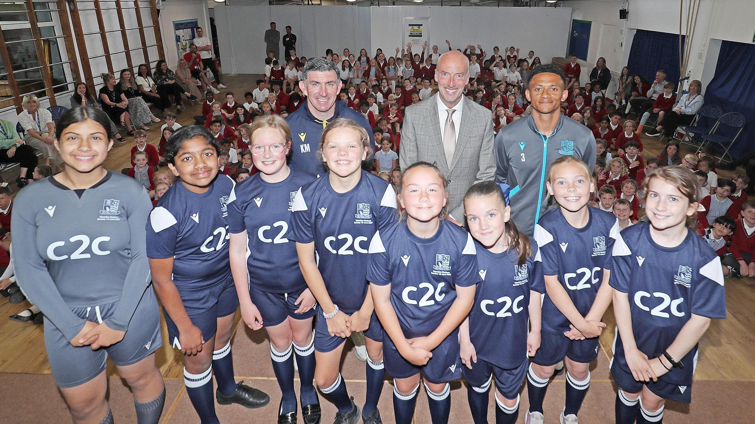 The girls, still wearing their blue kit, stand with their head teacher, Kevin Maher and Nathan Ralph on a stage inside their school hall. They are smiling and are in front of all their fellow school pupils, who are sitting on the floor.