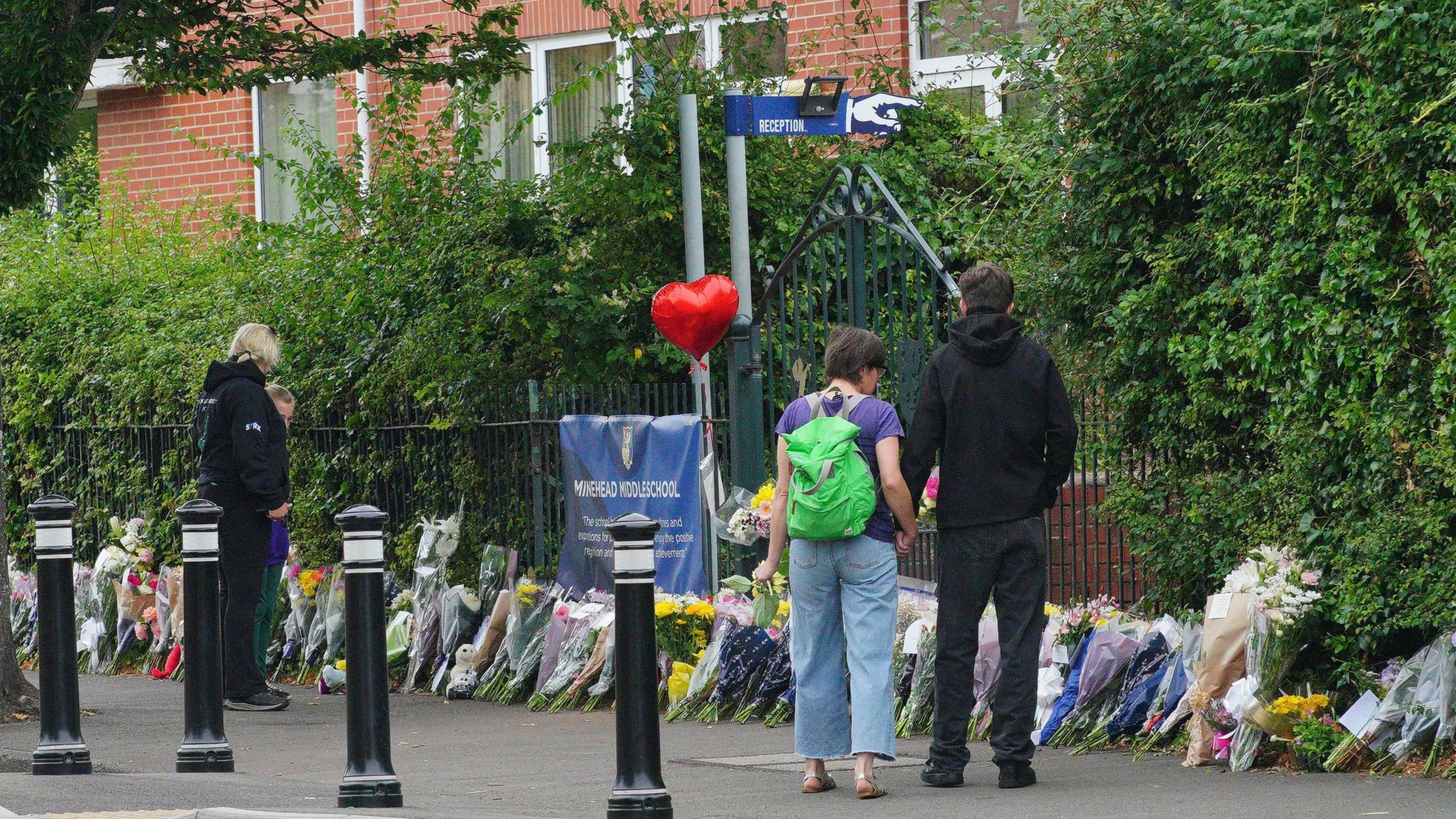 A few people look down at floral tributes placed outside Minehead Middle School. There are lots of bunches of flowers places along the pavement, next to the fence. 