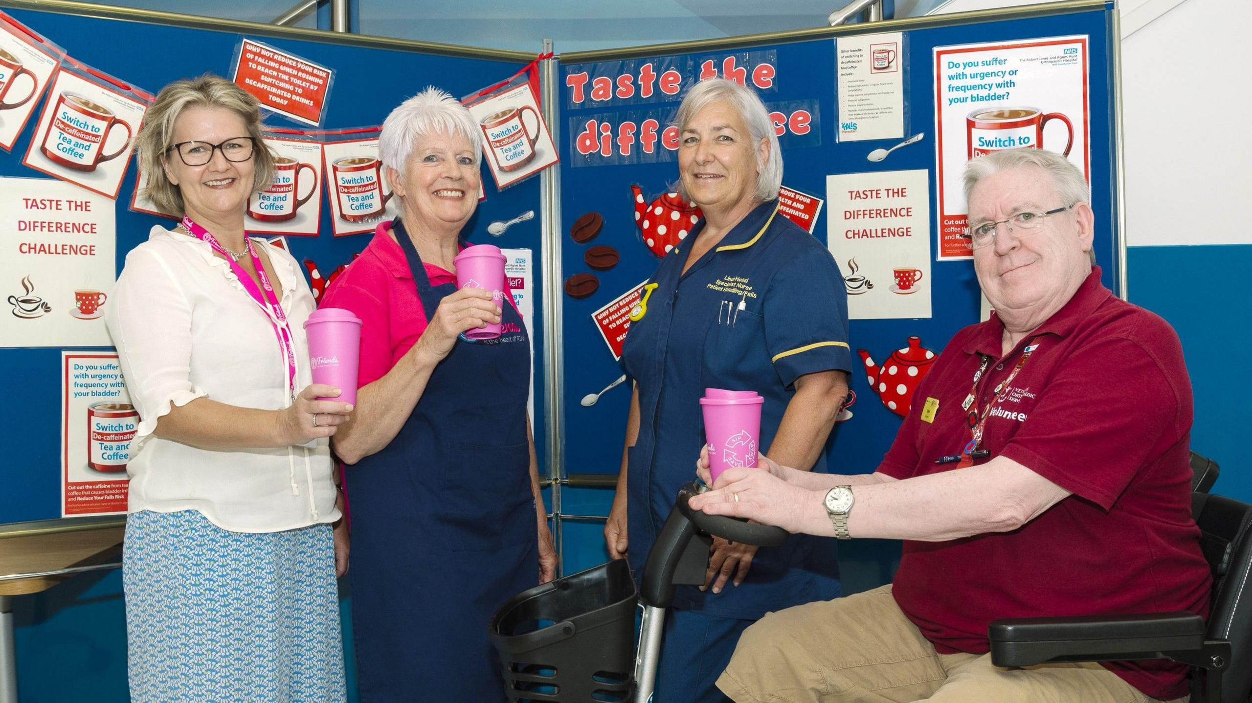 Three women are standing in front of a blue bulletin board with posters all over it. A sign on it reads "taste the difference". They are holding pink reusable cups. There is a man on the right hand side sitting in a motorised scooter, also holding a pink reusable cup