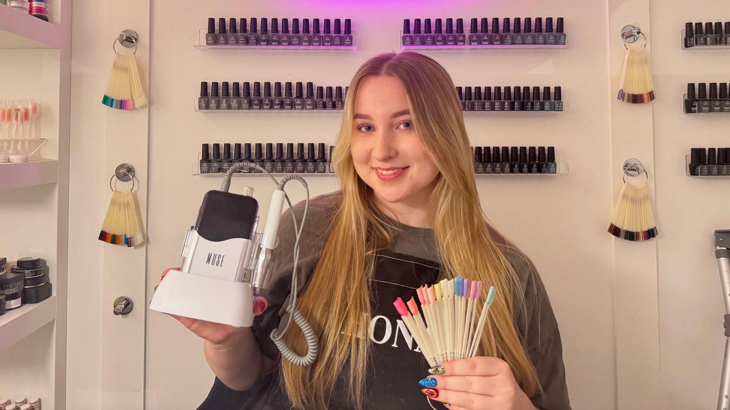 Faye Husband has long blonde hair and is wearing a grey jumper with a black apron over the top. She is holding an E-file in her left hand and nail colour samples in her right. She is standing in front of shelves of nail polish bottles in her home studio.
