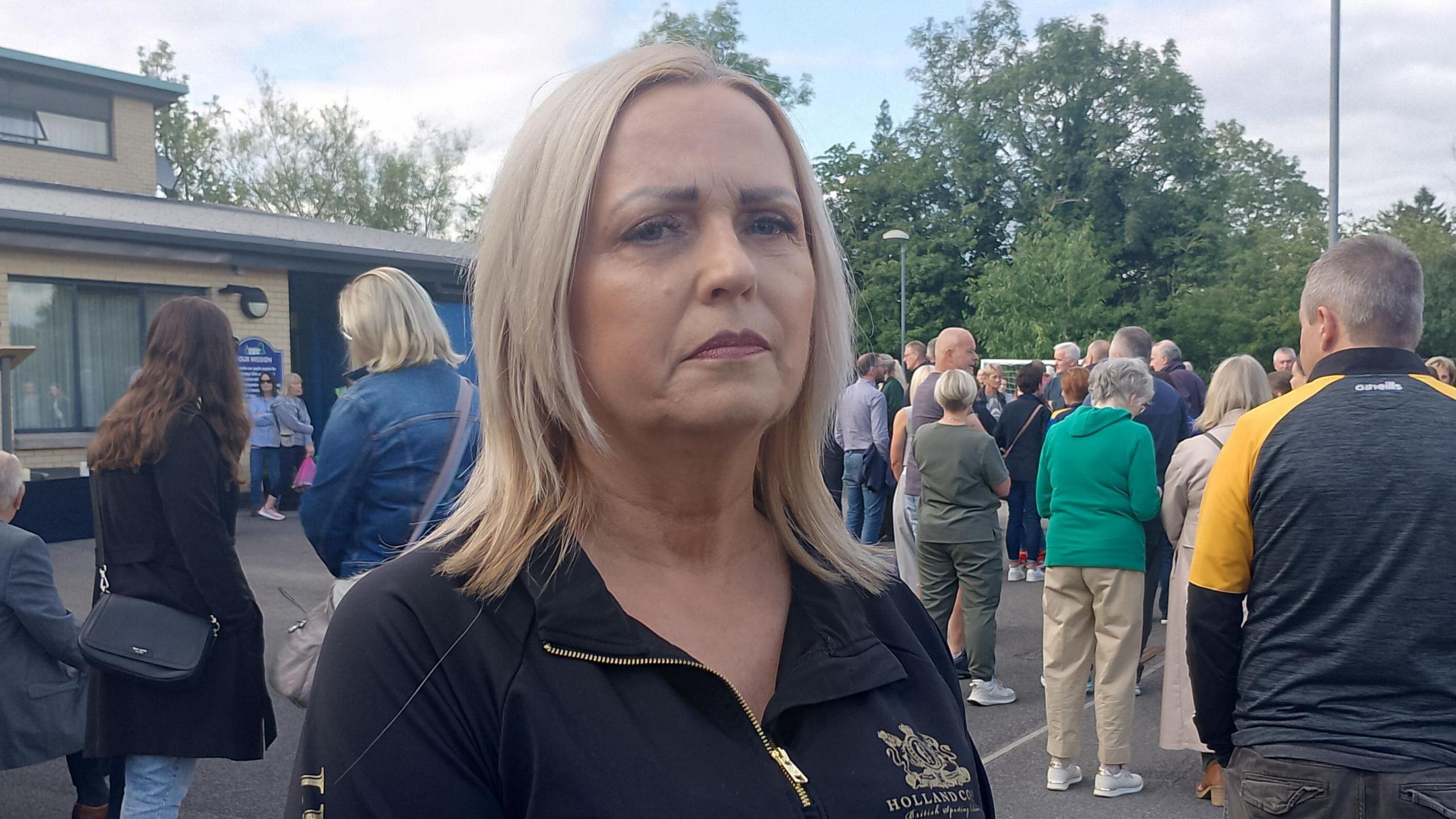 Anita has shoulder-length blonde hair. She is wearing a black zip up jacket.