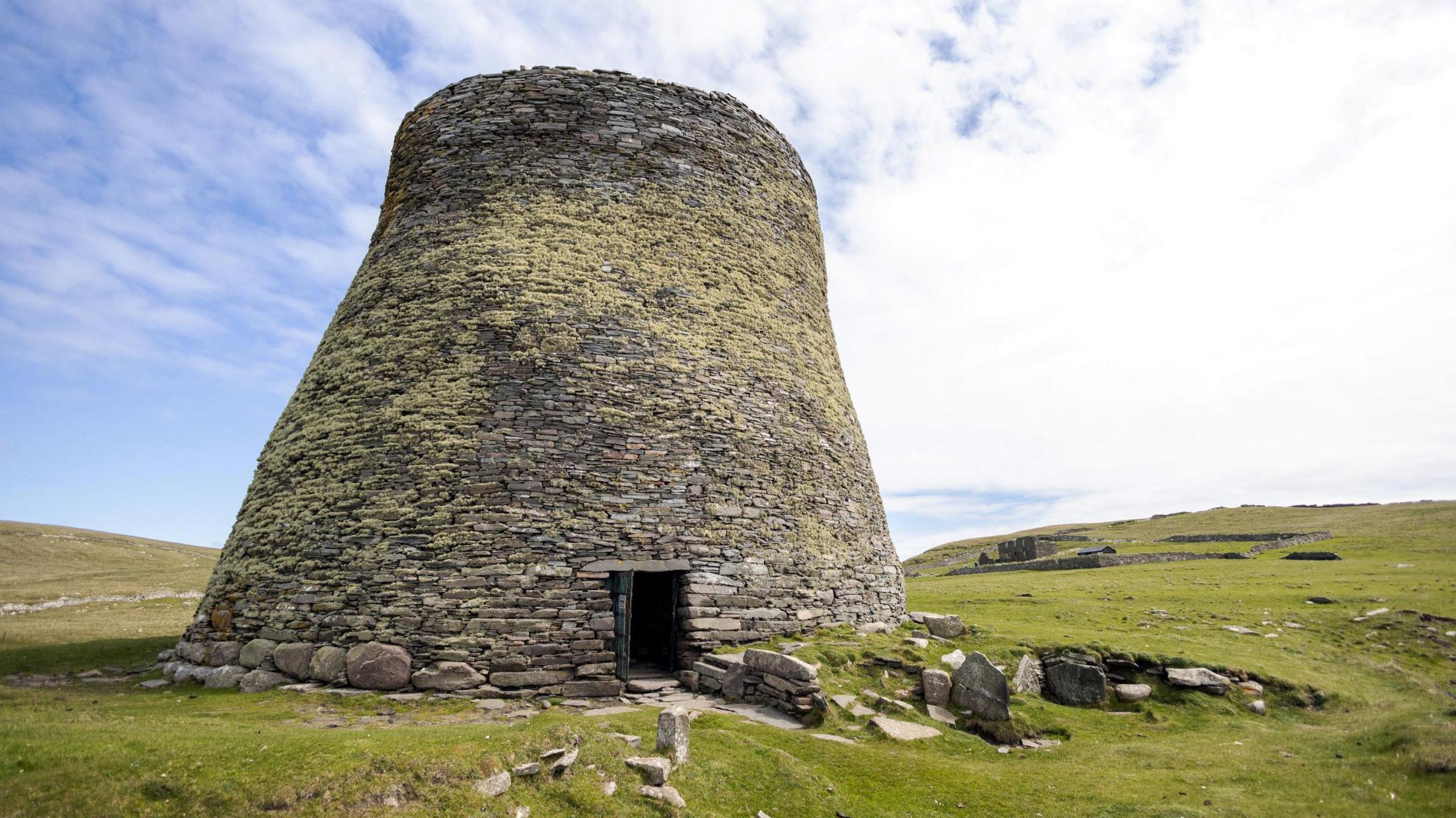 The broch is a tall, circular stone-built tower in a flat area of ground of closely-cropped grass. The walls of the tower are covered in light green lichen.