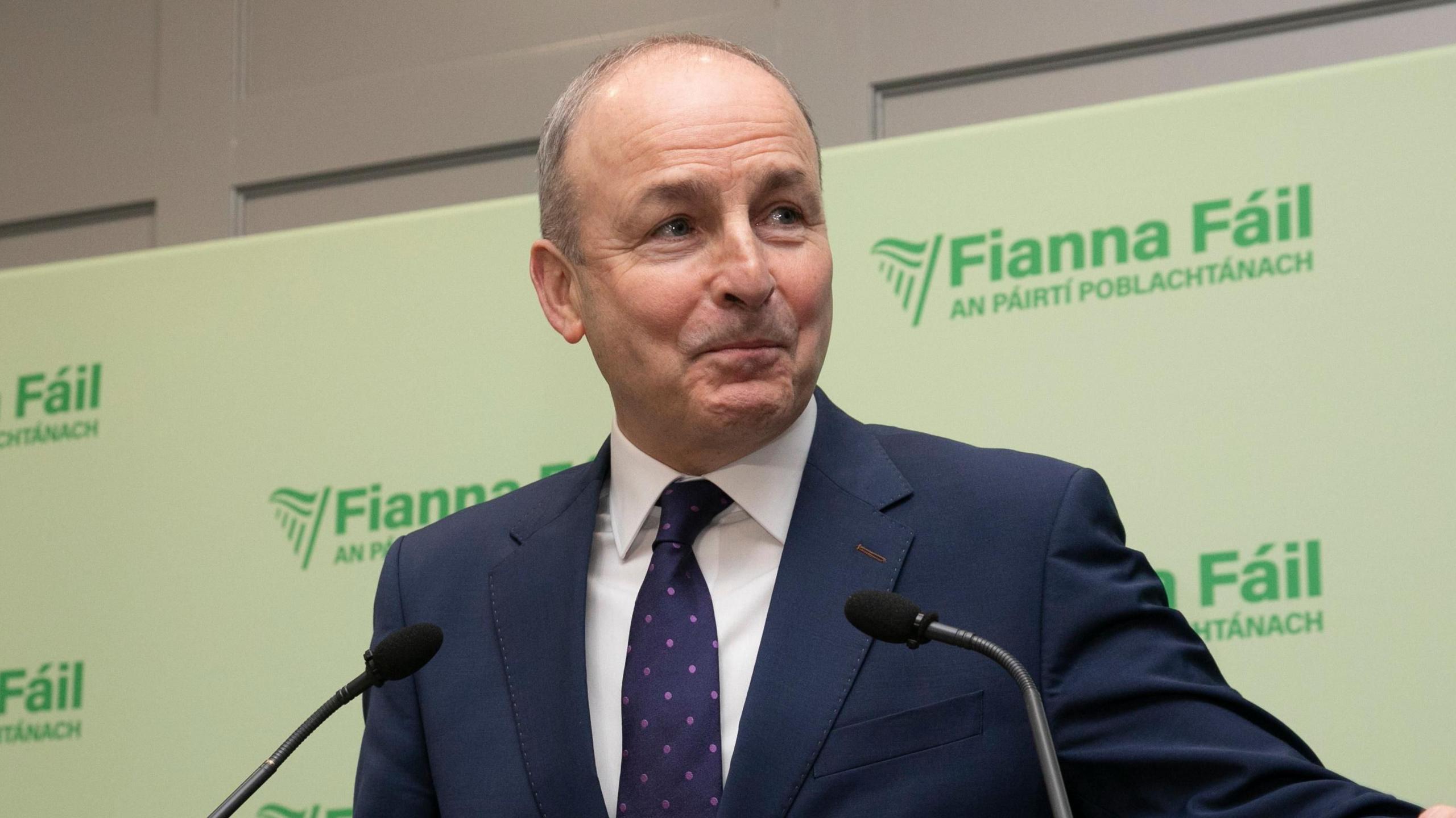 The Fianna Fáil leader, Micheál Martin, standing in front of a green Fianna Fáil background. He is wearing a navy suit jacket, white shirt and purple tie with pink dots on it. 