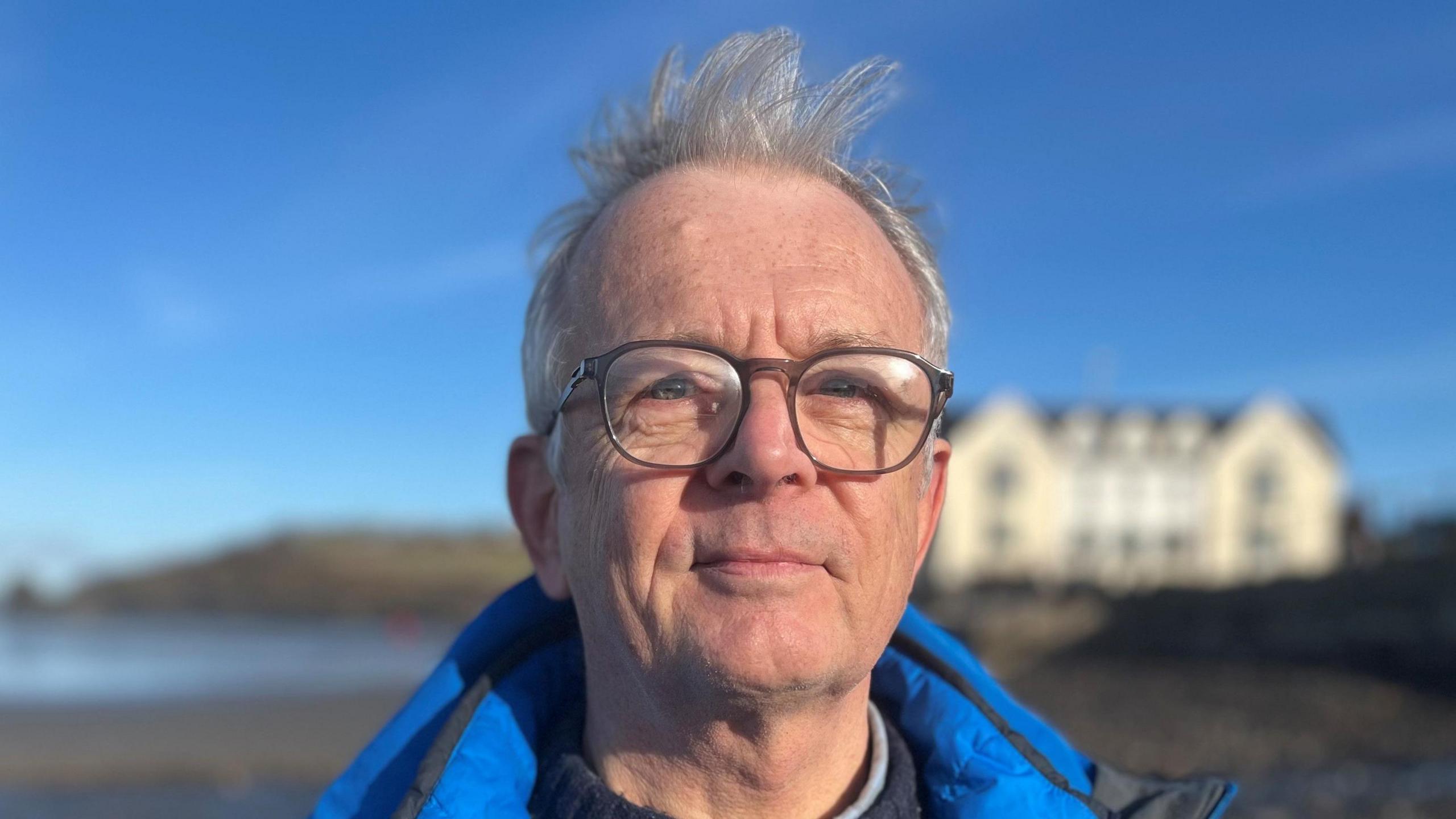 Andy Drumm faces the camera. He has grey hair and glasses and is wearing a blue coat. In the background can be seen the beach and a large white building, although out of focus.