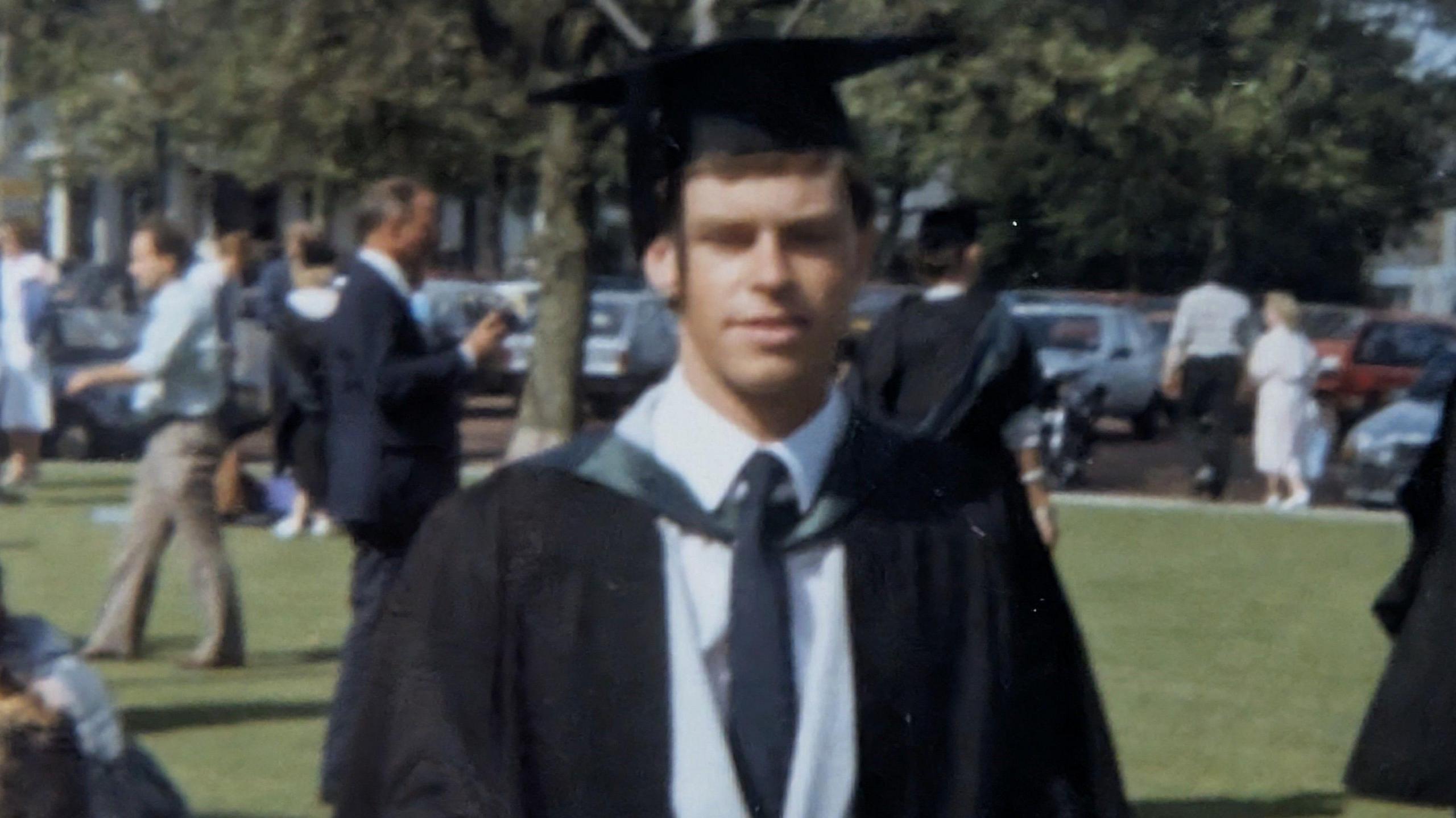 Alisdair Adams is seen as a young man on graduation day, wearing a light suit with a dark ceremonial robe and and mortarboard. He is standing in a field smiling with other students and families in the far distance.