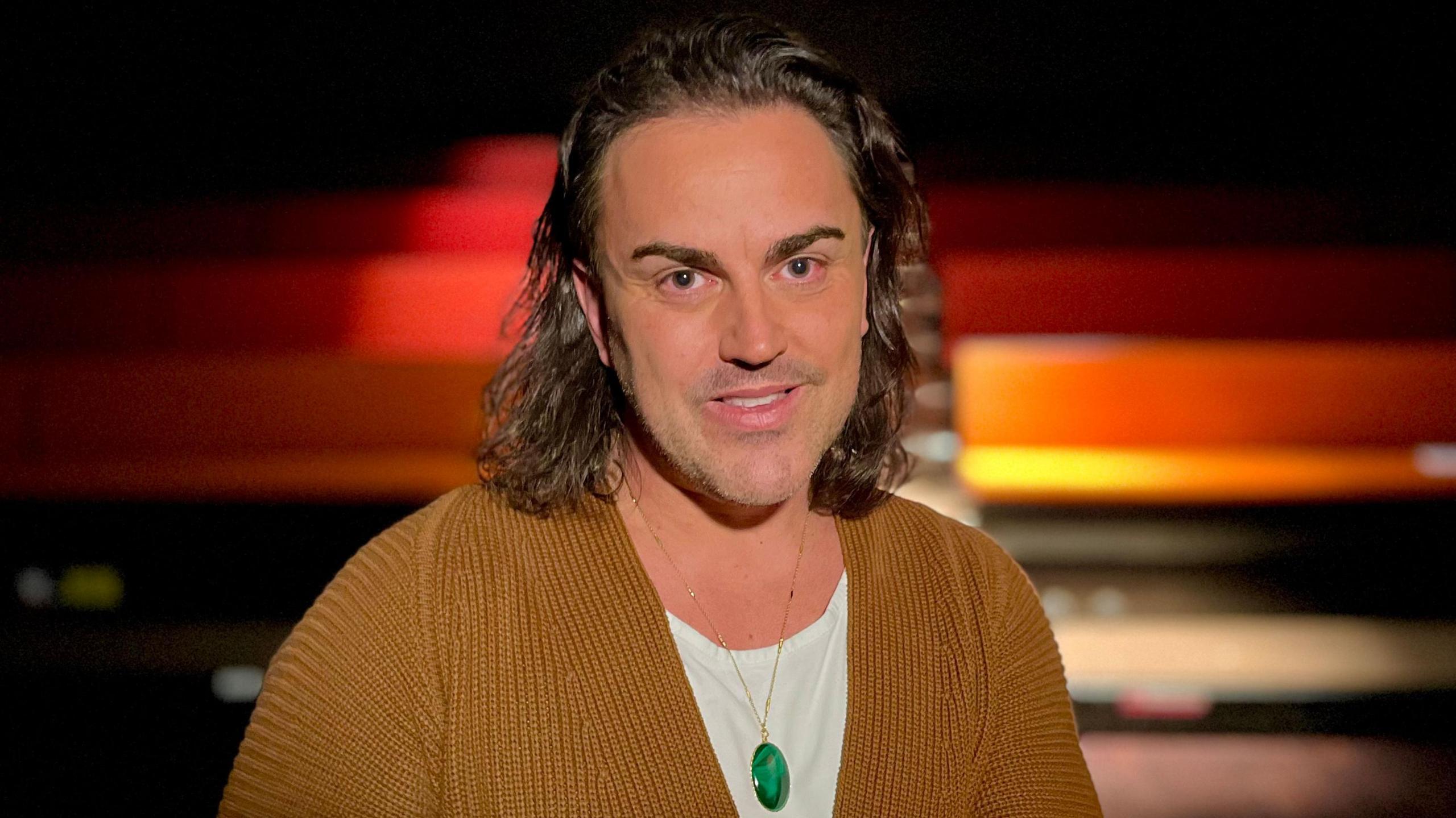 Stephen Crocker sits in a theatre he has long brown hair and is wearing a mustard cardigan, white t-shirt and green pendant necklace