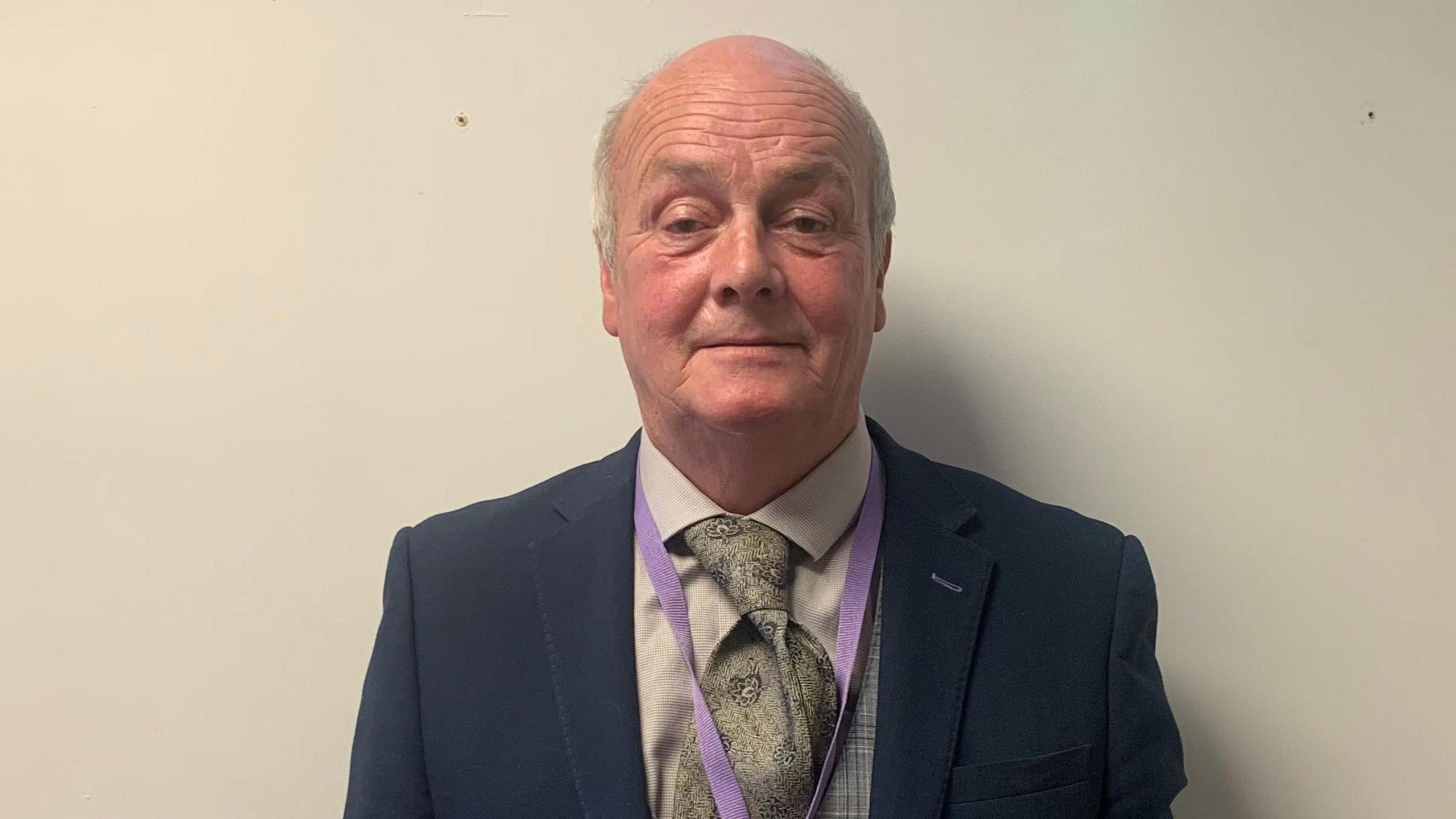 A bald man standing against a white wall. He is wearing a green tie and white shirt. He has a purple lanyard on and is looking at the camera.