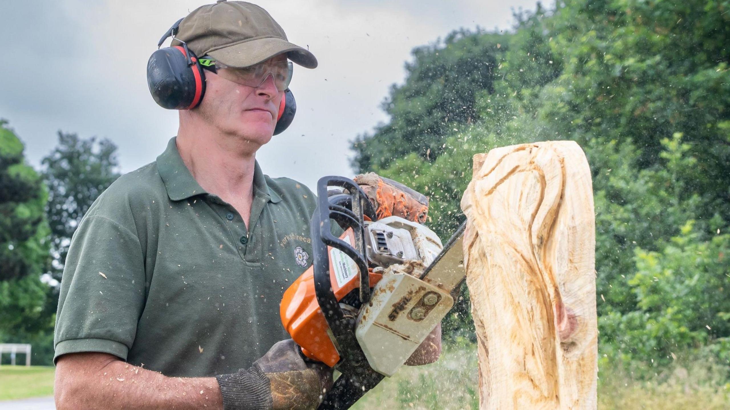 a man wearing ear defenders uses a chainsaw to carve a piece of wood