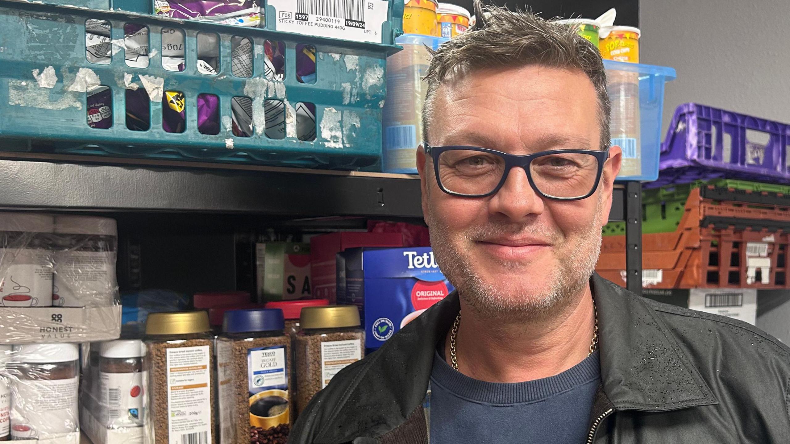 Tony Henderson stands in front of the shelves of goods in the food pantry. He has short brown hair, stubble and wears dark-rimmed glasses, a dark jacket and a blue T-shirt.