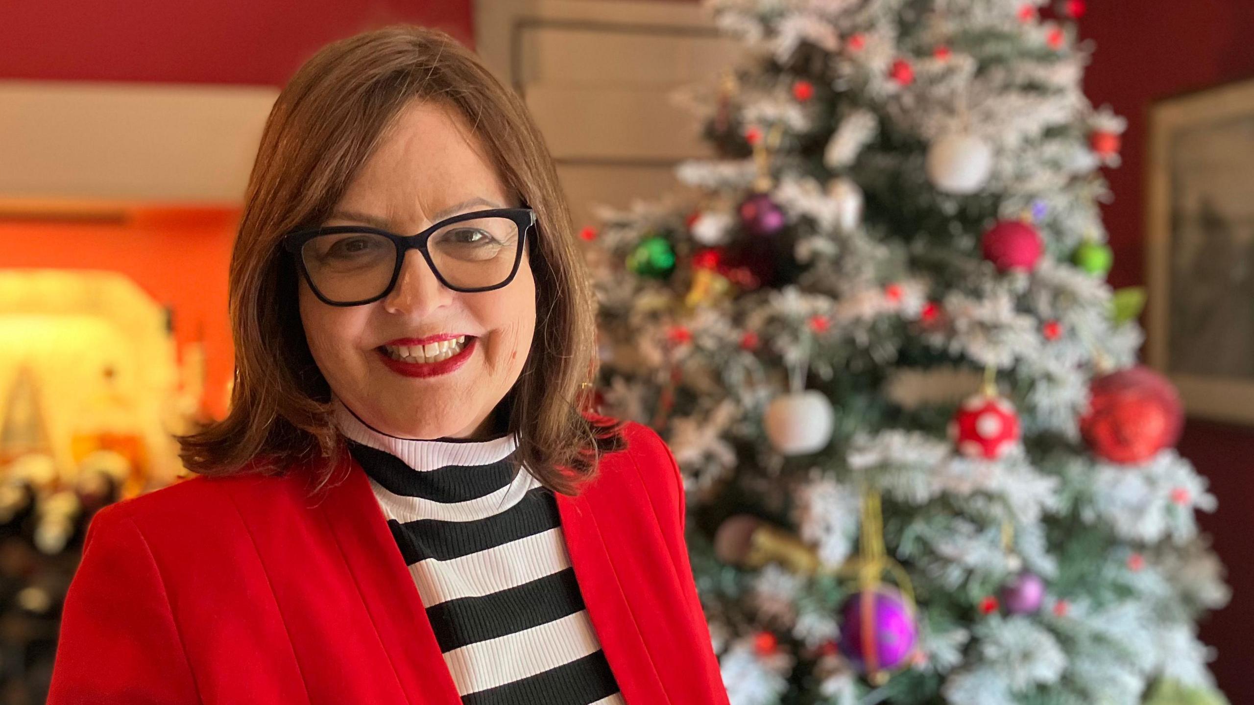 Jane Malyon smiling at the camera while standing in front of a Christmas tree in her living room. She is wearing a black and white striped jumper with a red jacket over the top.