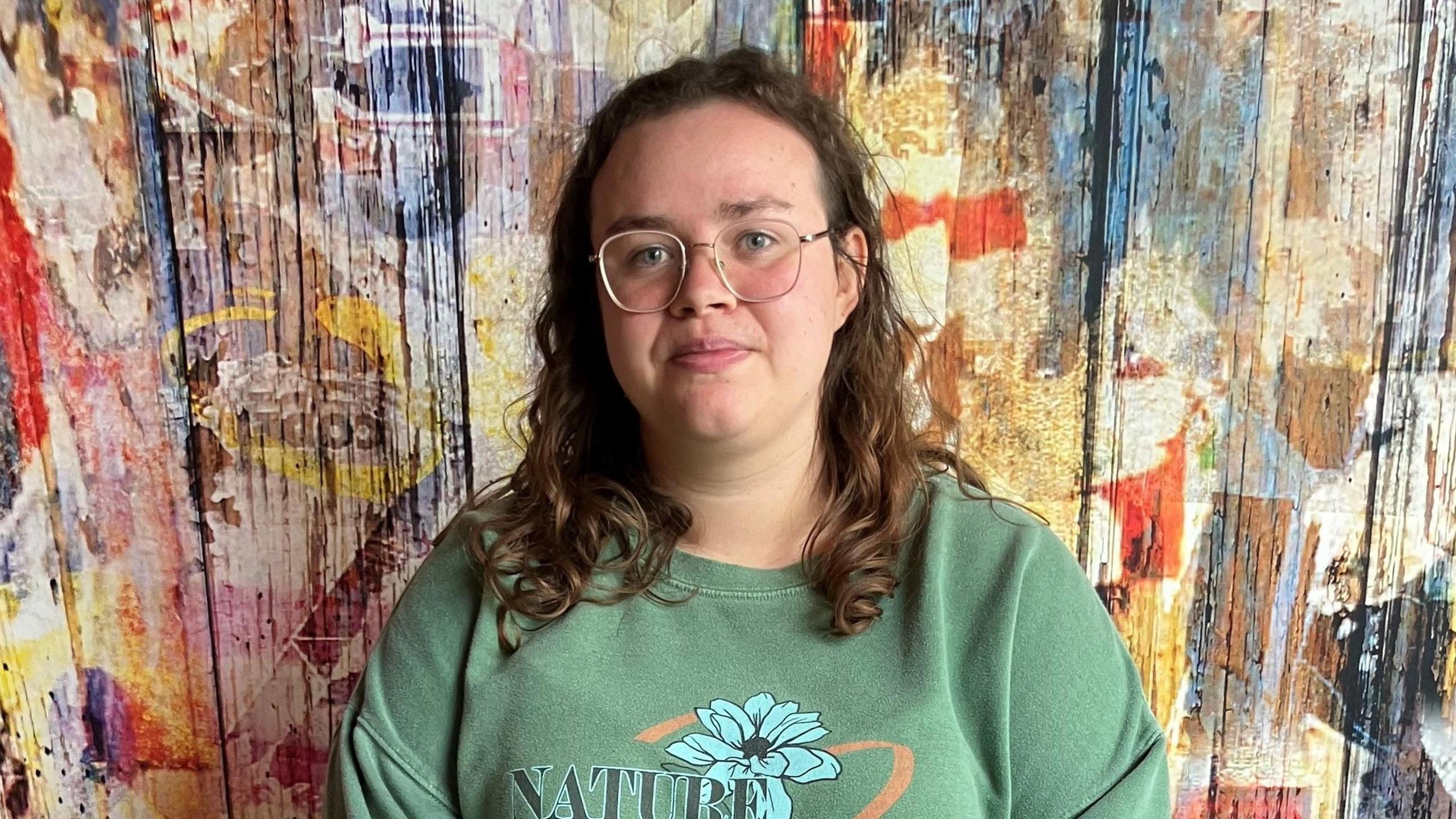 A young woman with curly long brown hair and wearing a green t-shirt with a blue flower and the word nature on the front. She is wearing gold round glasses and is standing in front of a multi-coloured wooden wall. 