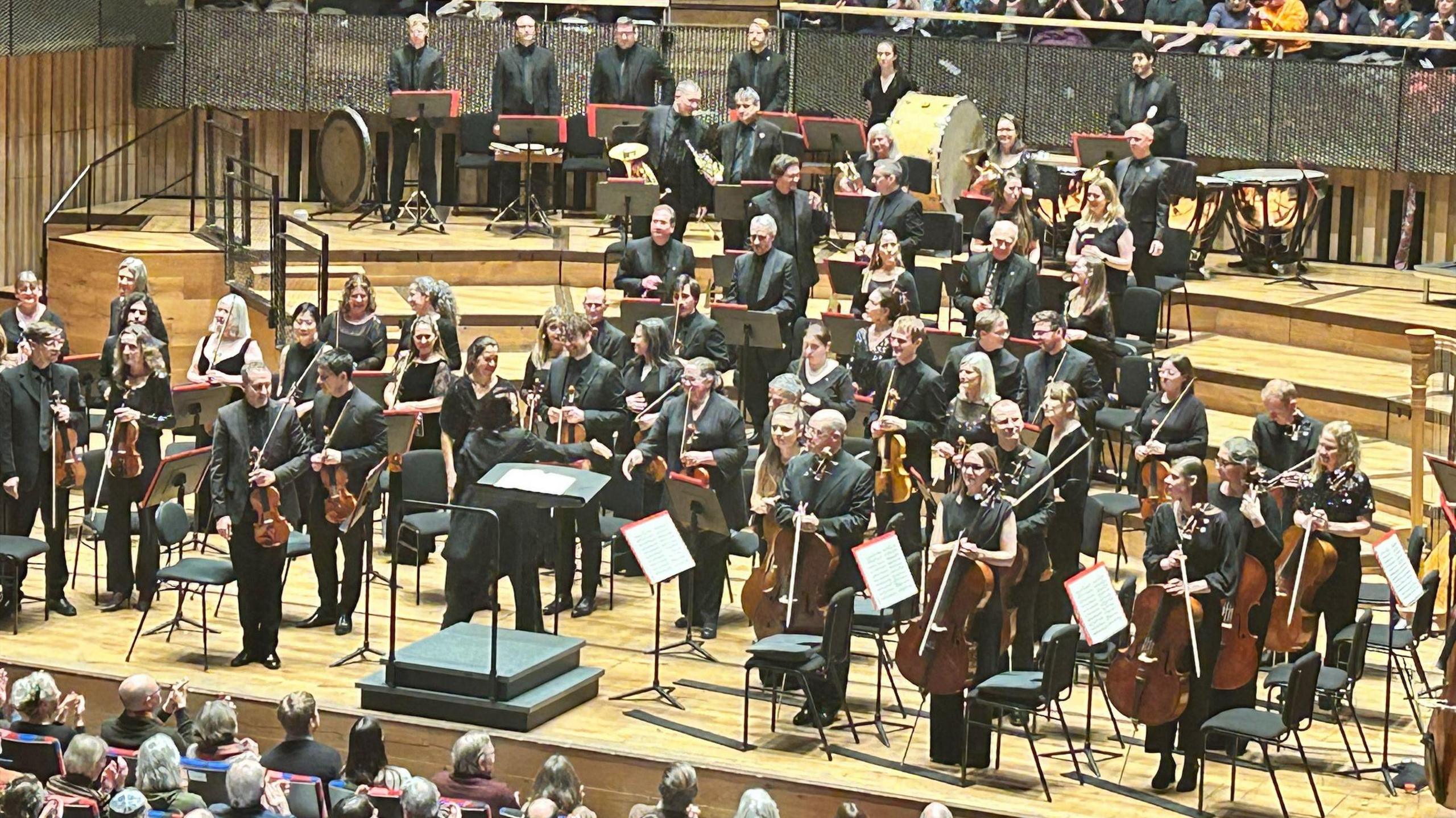 Members of the Bournemouth Symphony Orchestra perform on stage at the Bristol Beacon. The picture is taken from the back of the auditorium and the musicians, all dressed in black, are facing the audience and standing