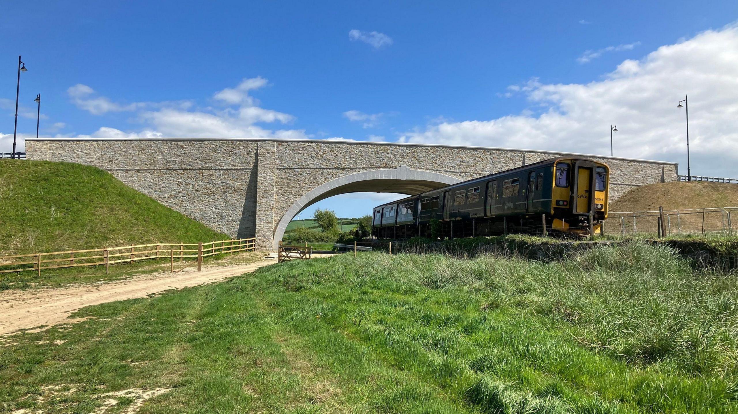 A train going under the new bridge. The bridge is clad in granite. There is grass and new wooden fencing near the train track. There are four lamp posts on the road leading across the bridge.