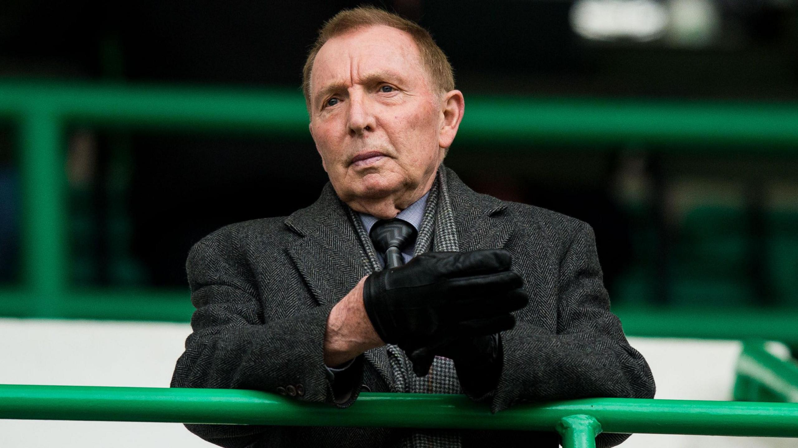 Sir Tom Farmer standing in a football stand. he is wearing a greay coat and grey shirt and tie