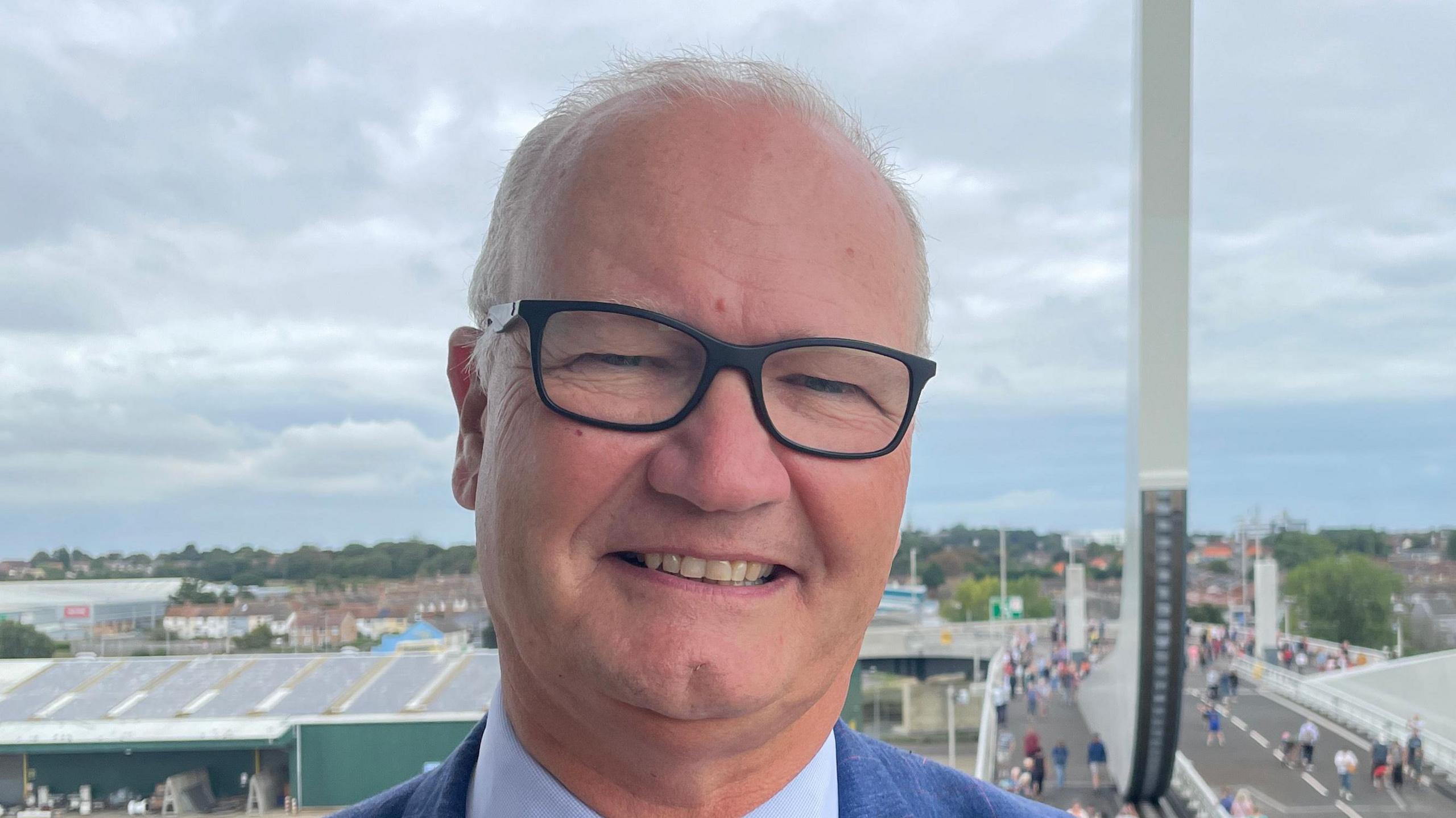 Matthew Hicks smiles at the camera. He has thin grey hair, wears glasses as well as a blue suit with a lighter blue shirt underneath. Behind him the Gull Wing Bridge in Lowestoft can be seen.