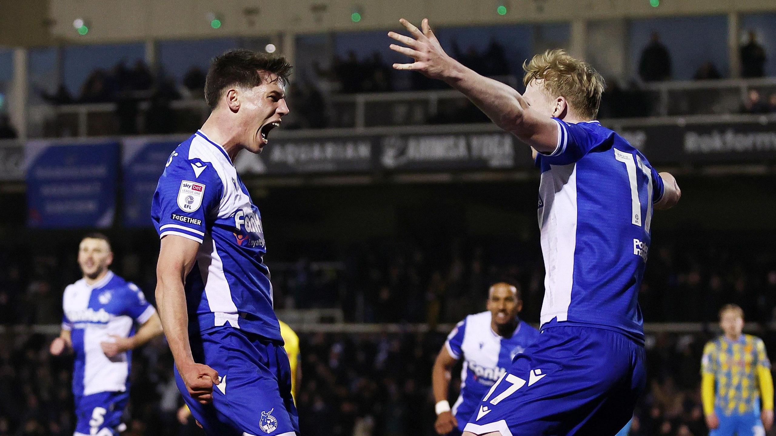 Two Bristol Rovers players, both wearing the club's traditional blue and white quartered kits, celebrate a late goal against Stockport County at the Memorial Stadium. One of the stadium stands is visible in the background