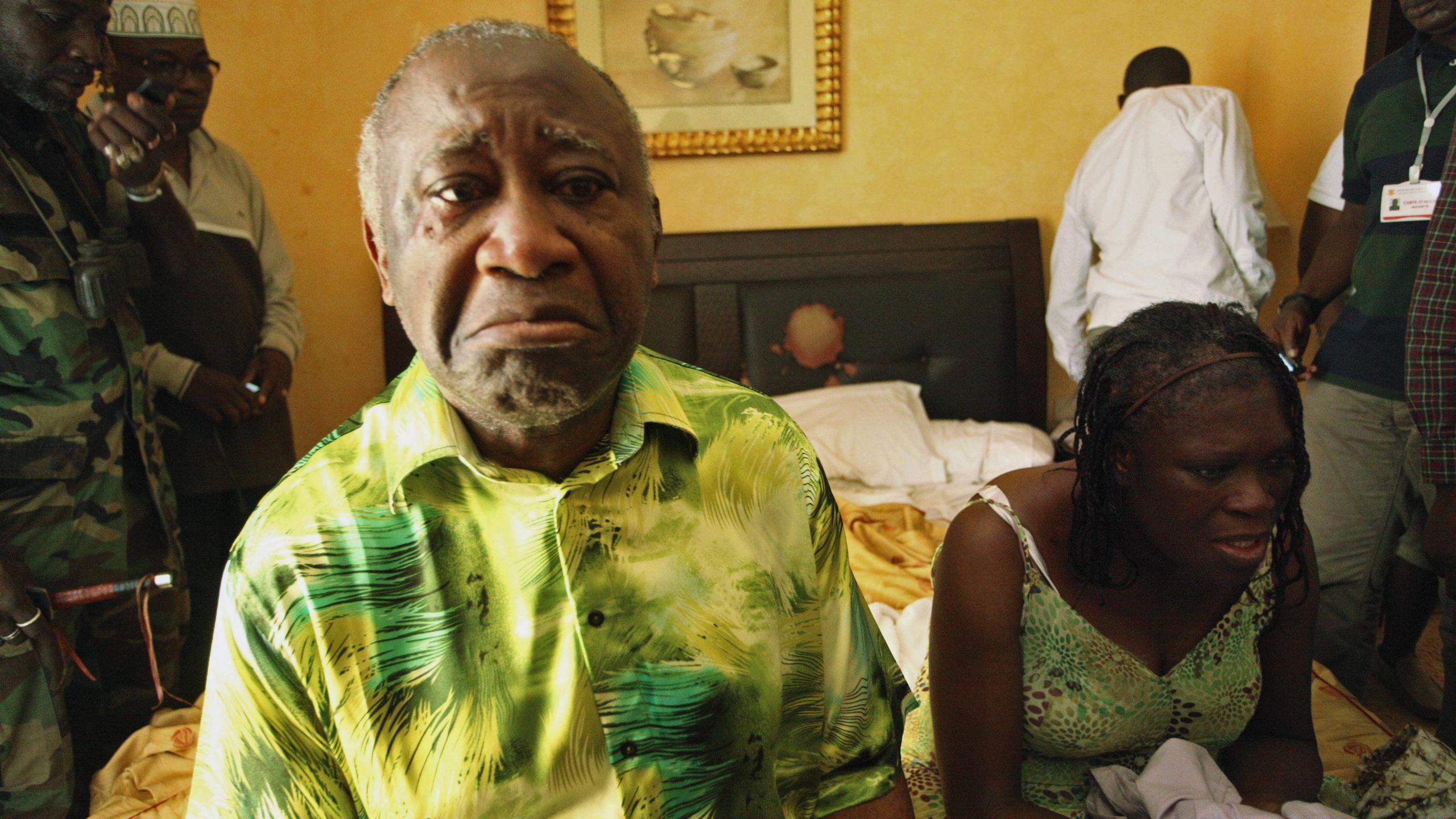 Laurent Gbagbo and Simone Gbagbo sit on the edge of a bed, with crestfallen expressions on their faces. Figures, including a man wearing a military uniform, stand behind them.