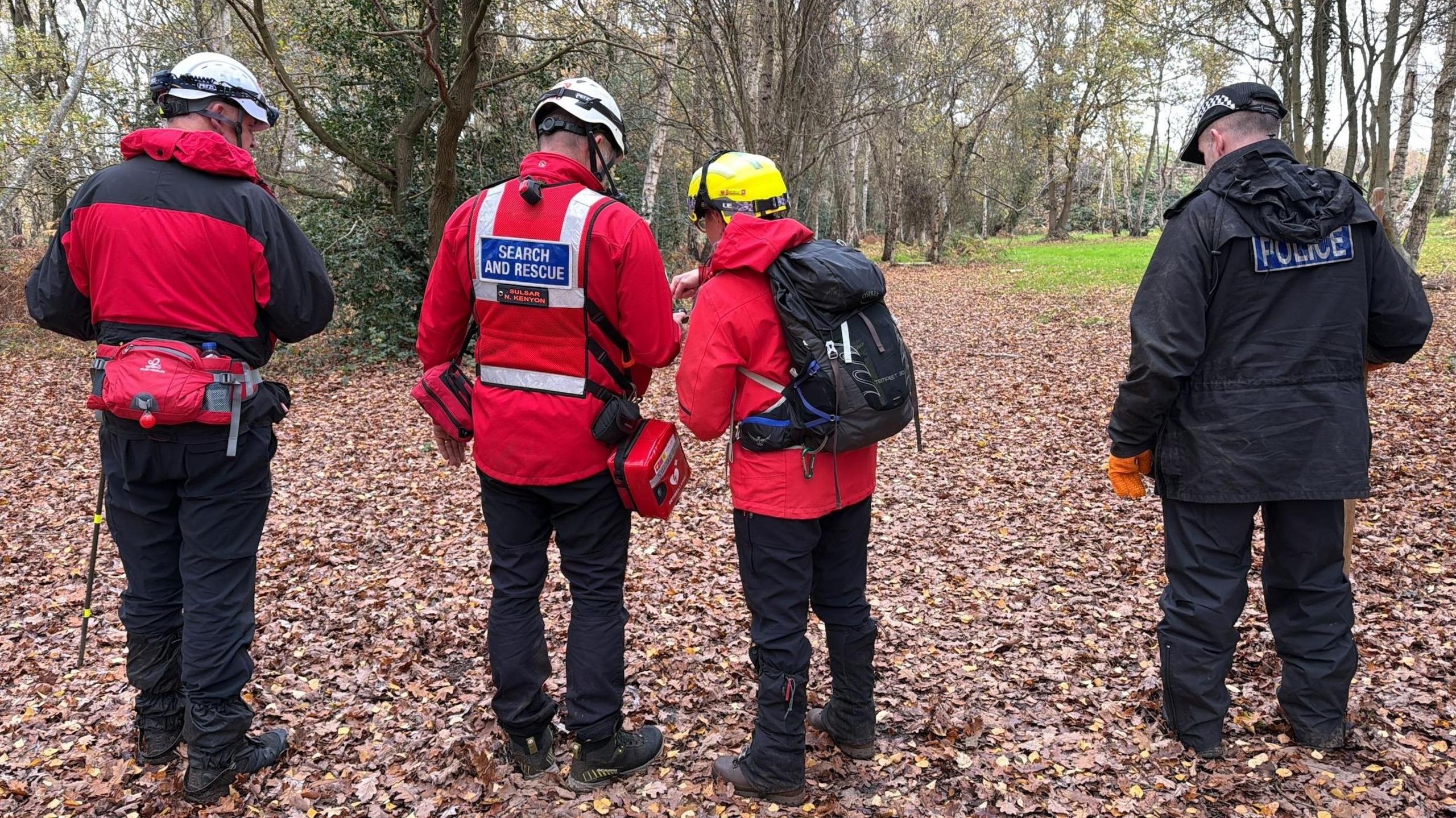 Four people are standing in a woodland which is covered in brown leaves on the floor. The three people on the left of the image have red coats and hard hats on and a person on the right is wearing all black with a POLICE logo on his coat.