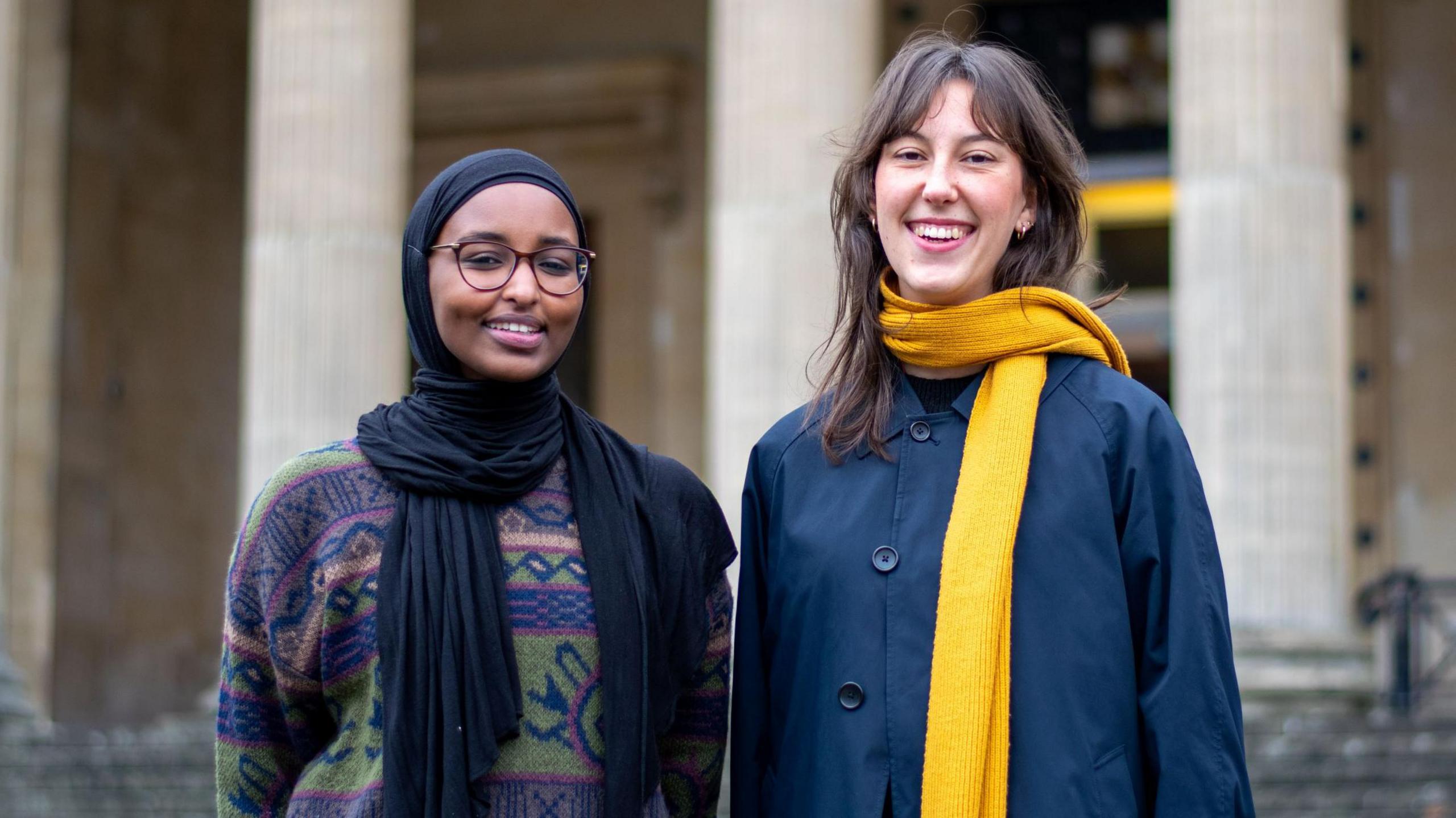 Two Bristol university students, both female, stand outside the Victoria Rooms in Bristol, both smiling at the camera. One is wearing a traditional headcovering while the other is wearing a blue coat and yellow scarf