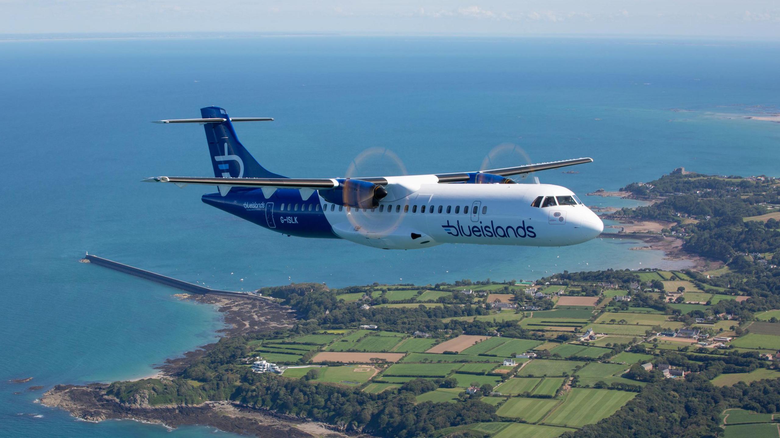 A plane in the sky flying over land. There is green land on the right and sea on the left.