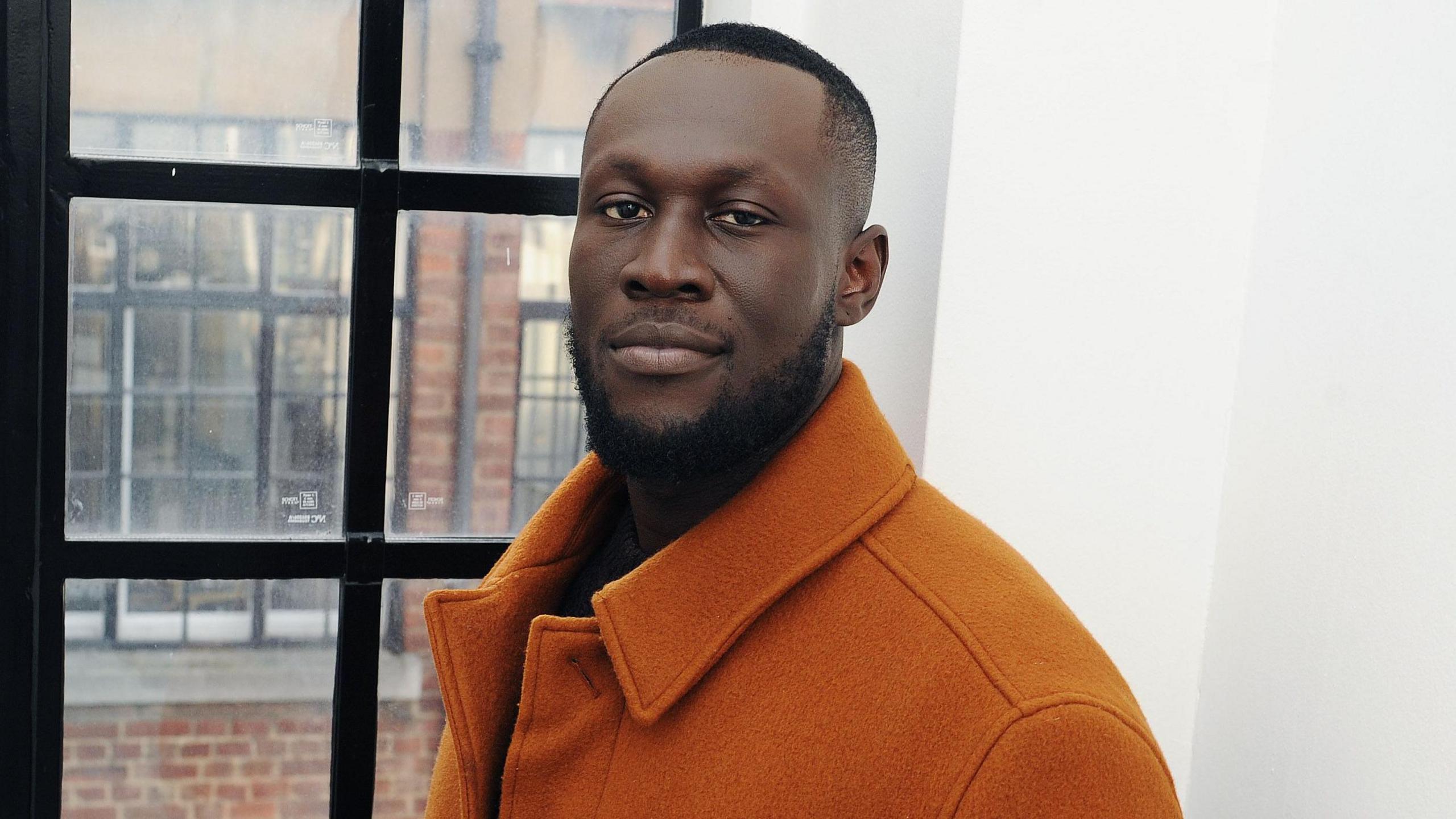 Stormzy looking into the camera, with a slight smile on his face, standing to the side in front of a window, wearing a burnt orange coat. 