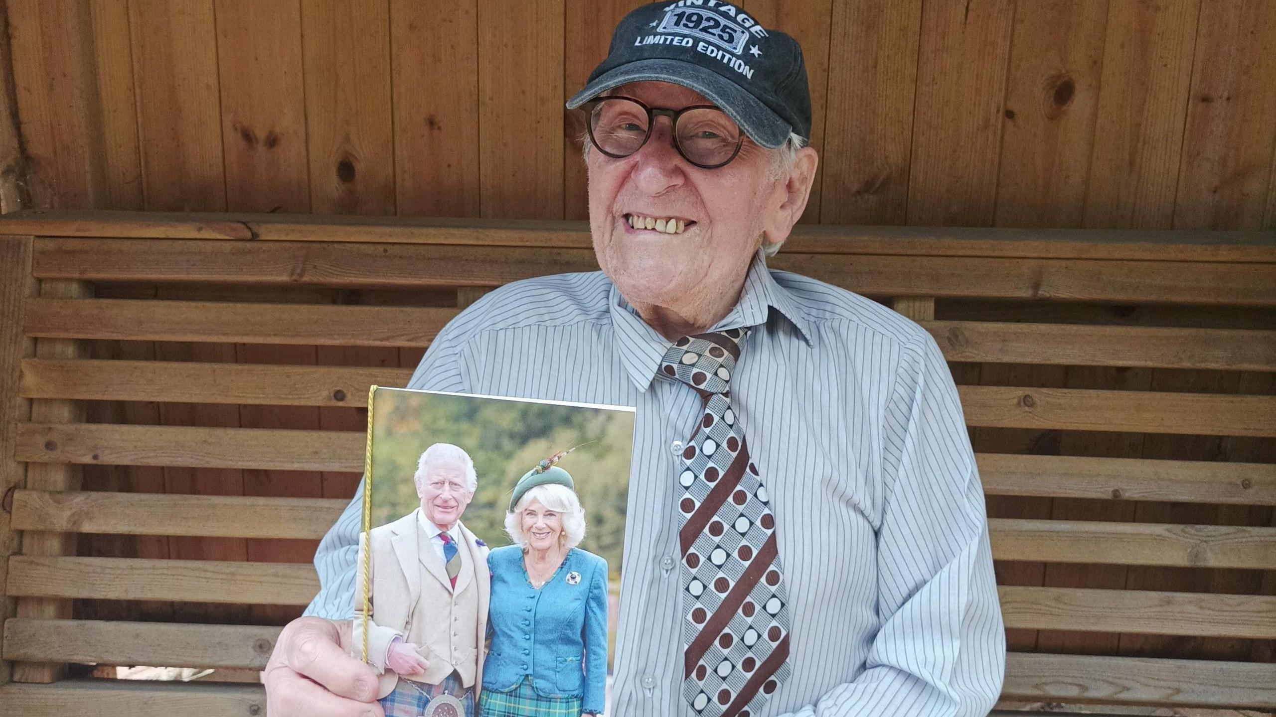 Bill sits in a wooden hut and is wearing a blue and white striped shirt, and grey, white and maroon tie with stripes and dots on it, and dark grey baseball cap with a logo and some writing on it, one bit of which says 1925. He is holding up a photo of the King with his wife, Queen Camilla towards the camera.