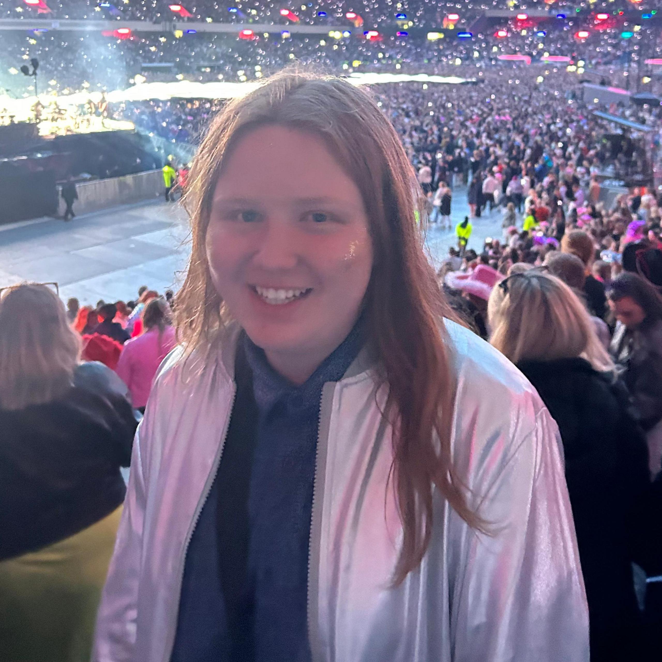 A boy with long hair wearing a white jacket in a stadium. There is a big crowd in the background and coloured lights. 