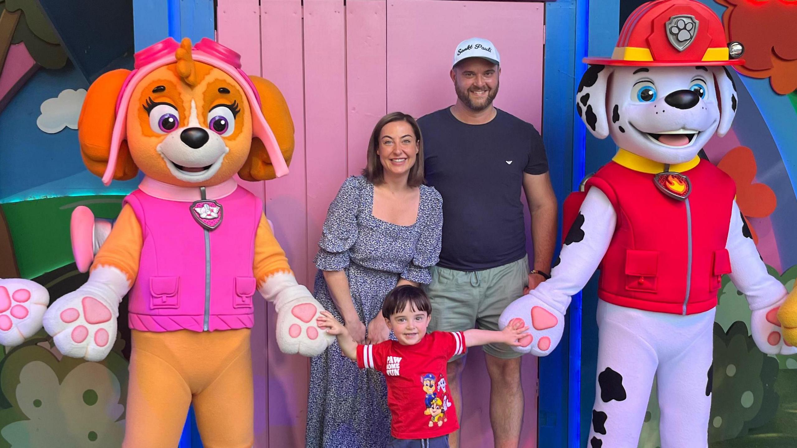 A woman with bobbed brown hair, wearing a long black and white dress, stood next to a man with a white baseball cap, blue t shirt and khaki shorts. In front of them is a young brunette boy in a red Paw Patrol character t shirt. They are stood in front of a bright pink door, and on eother side of the boy are people dressed up as dogs from Paw Patrol tv series.