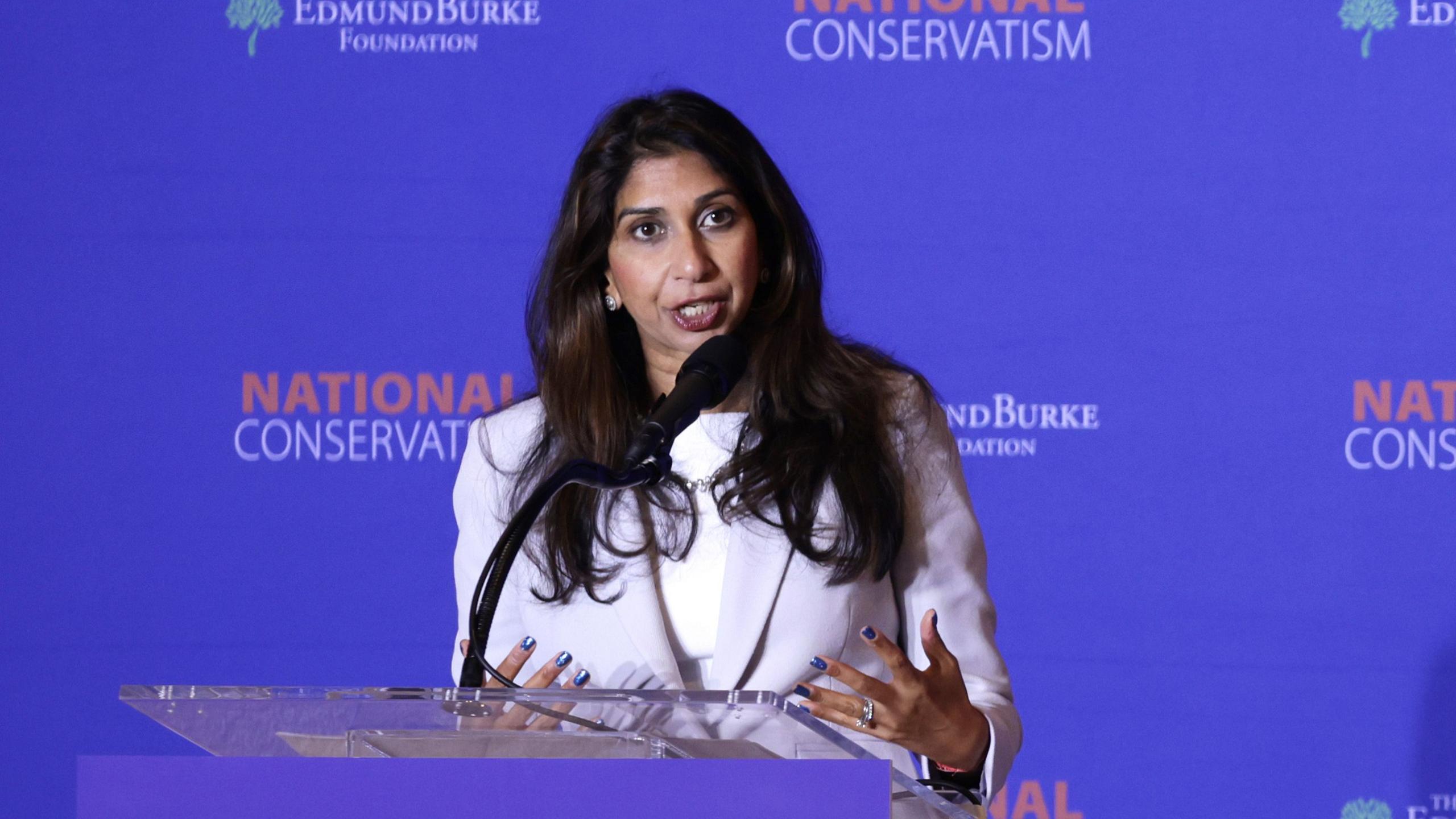 Suella Braverman standing at a lectern with a microphone. There is a blue background which reads 'National Conservatives' 