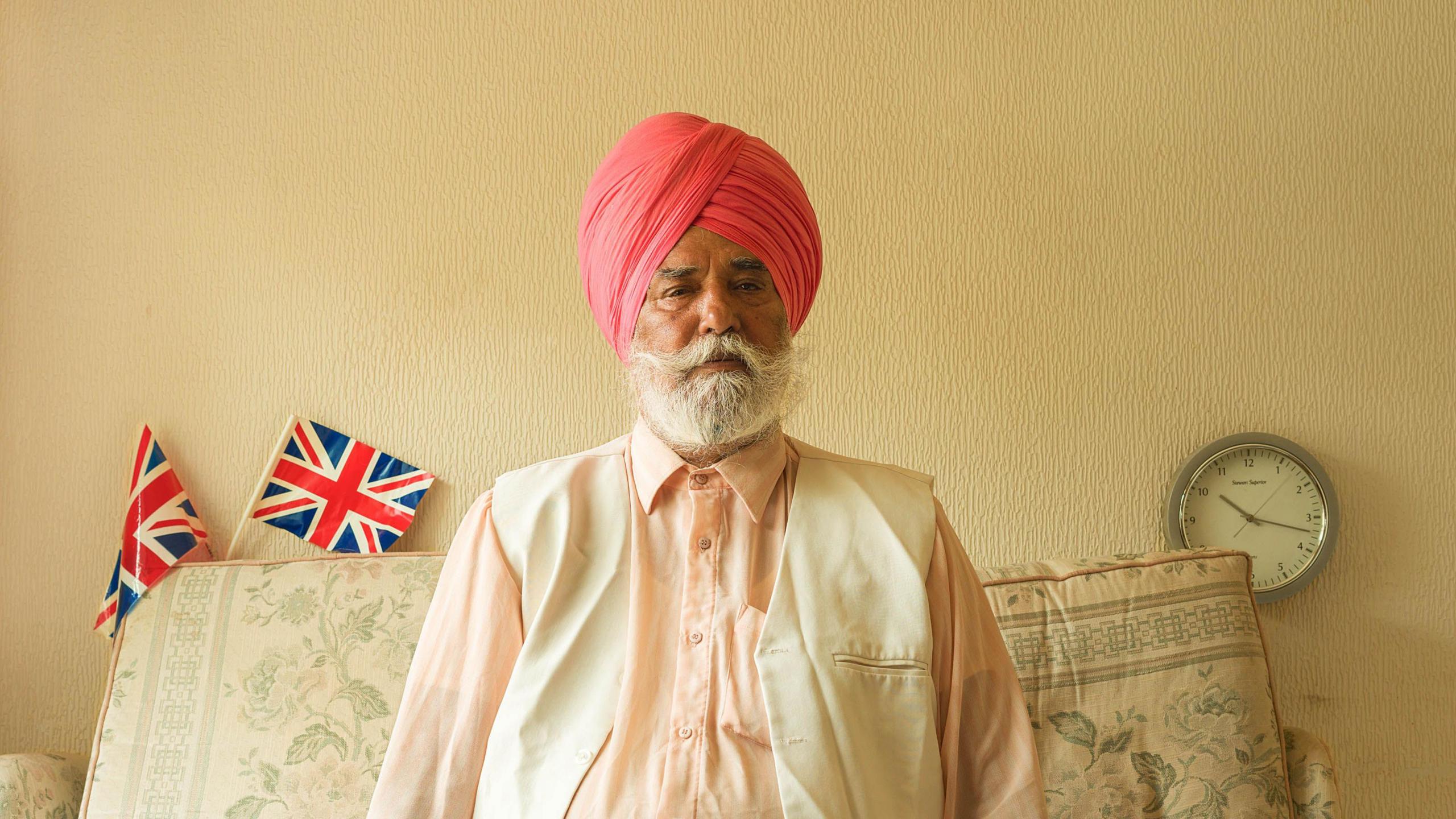 A man sits on his sofa wearing a light waistcoat and pink turban with union jack flags arranged on the back of the sofa