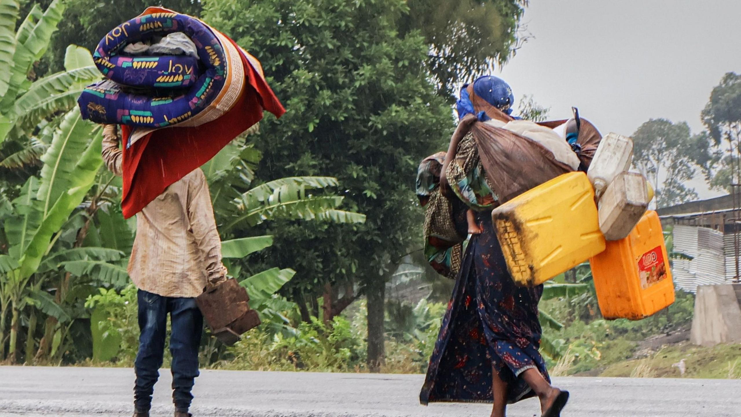 A man and woman carry belongings such as a mattress and water containers.