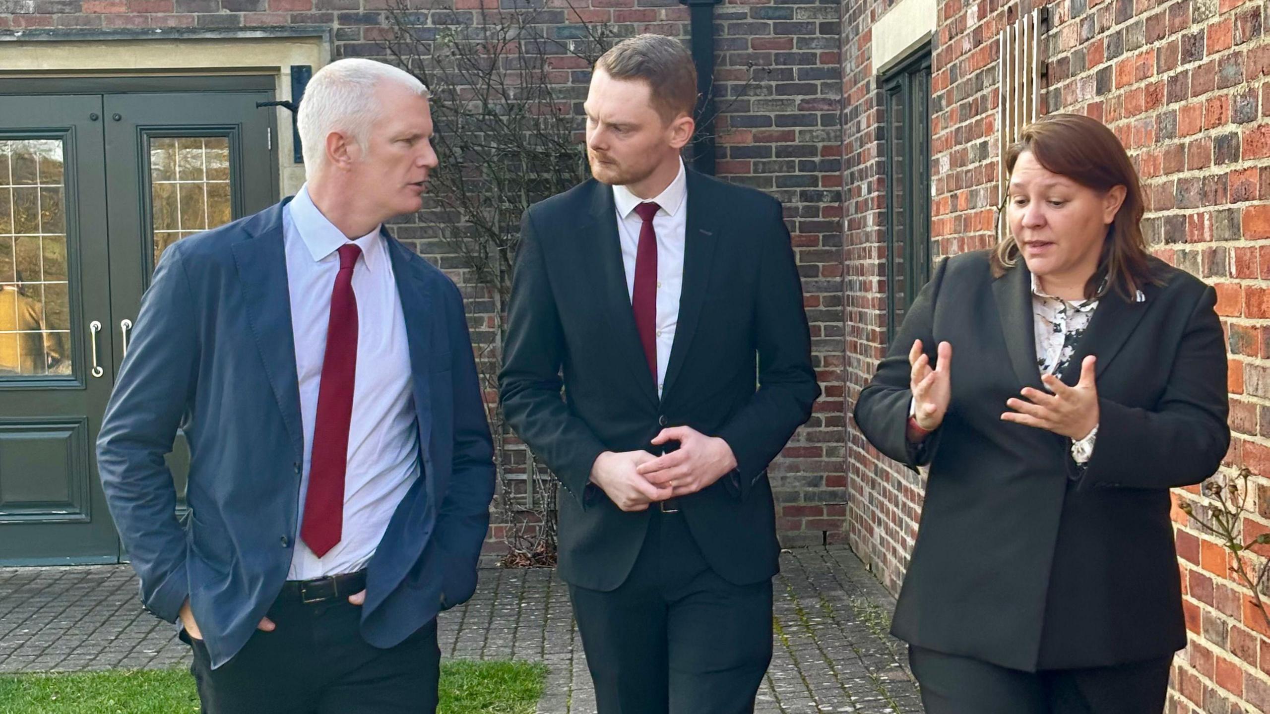 Alistair Carns, Luke Myer and Anna Turley talking outside. Carns has grey, short hair and is wearing a blue suit jacket with a red tie. His hands are in his pockets. To the right is Myer who has brown hair and is wearing a dark suit with a red tie. He's looking at Carns. On the right is Turley who has long brown hair and is wearing a dark suit. She's gesticulating with her hands.