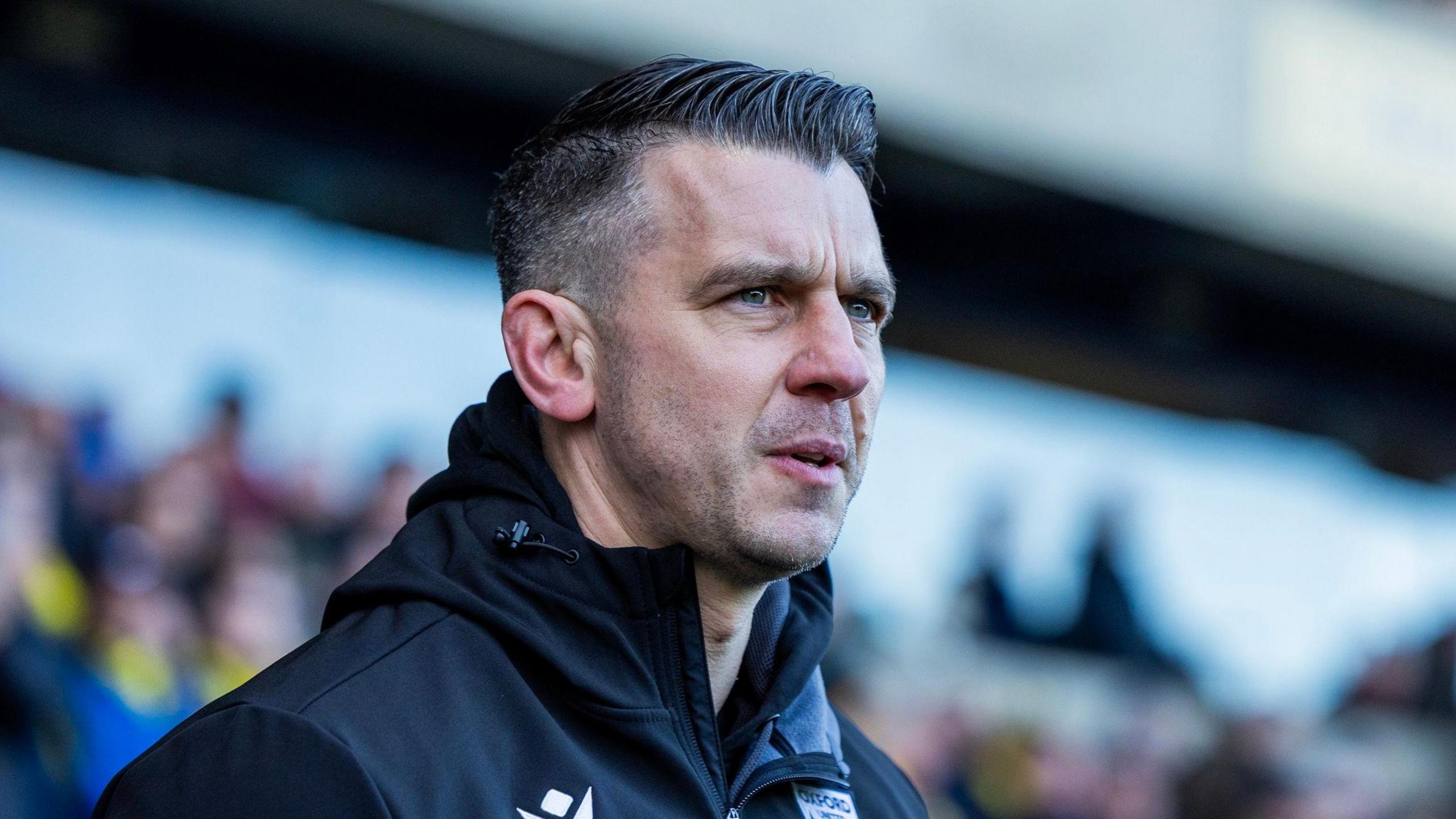 A close up of Matt Bloomfield wearing a black, hooded jacket with a blurred image of Oxford United fans in the background