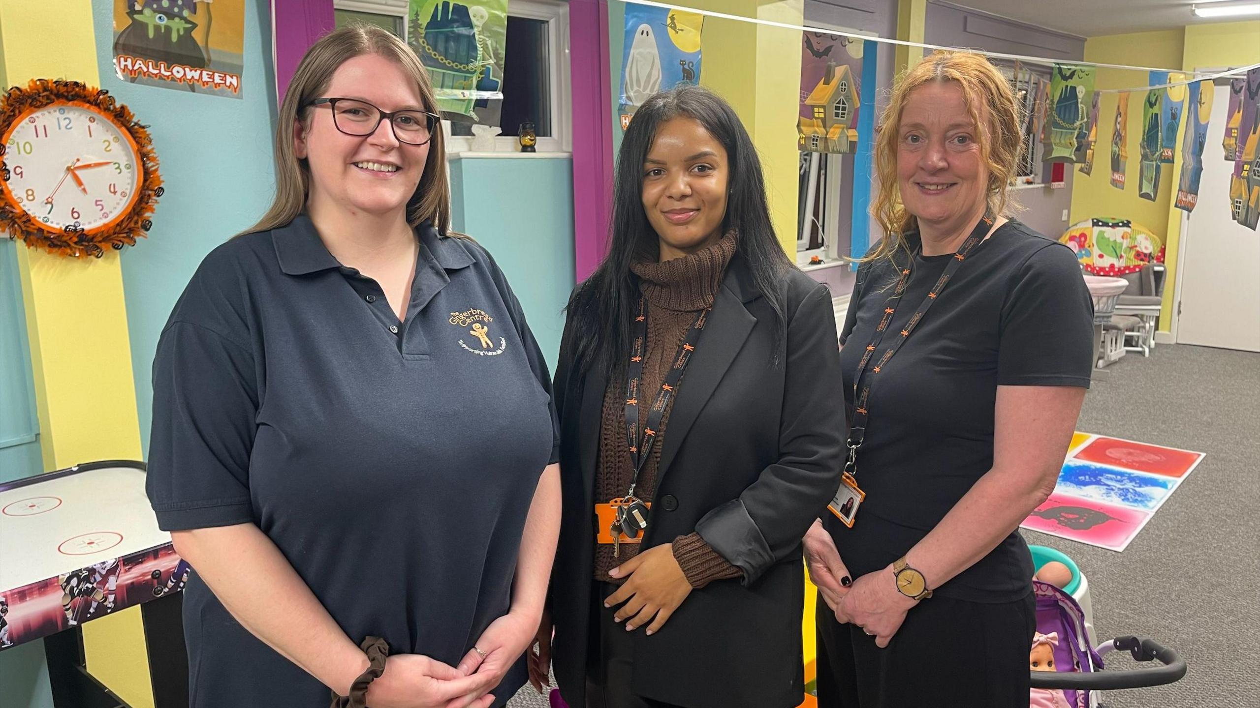 Three women smile at the camera. They stand in front of a wall painted mainly blue with other colours like yellow in broad stripes on it. On the wall are pictures done by children and a brightly coloured clock. The left hand woman has glasses and dark brown hair past her shoulders and wears a dark blue polo shirt. The women in the middle has long black hair past her shoulders and wears a brown jumper with a black jacket over it - a lanyard is around her neck. The woman on the right has shorter, light red hair and wears a black T-shirt.