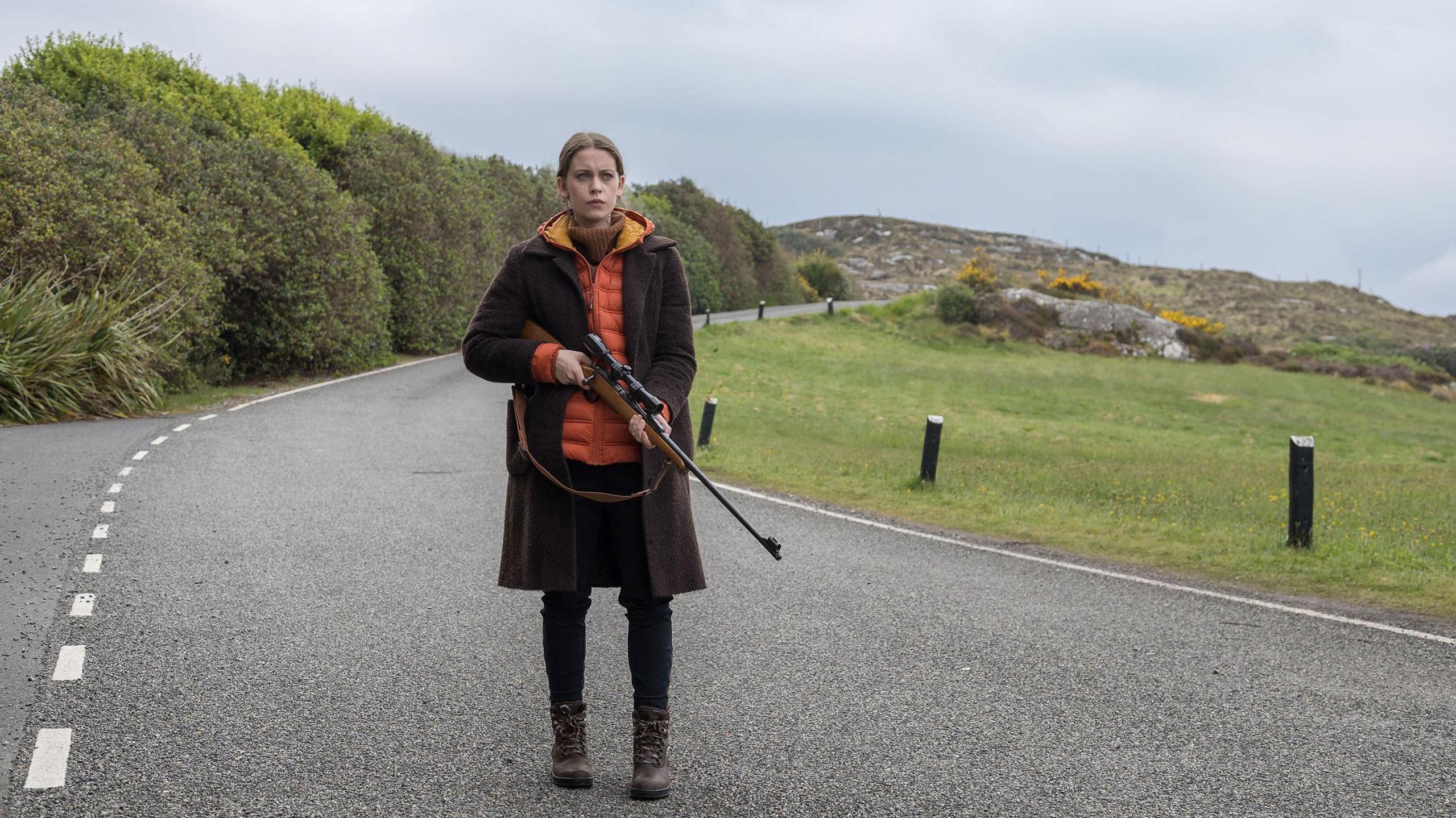 Star Sorcha Groundsell as Kat in An t-Eilean (The Island) - she is walking along a country road while holding a rifle. She has auburn hair and a brown and orange jacket on.