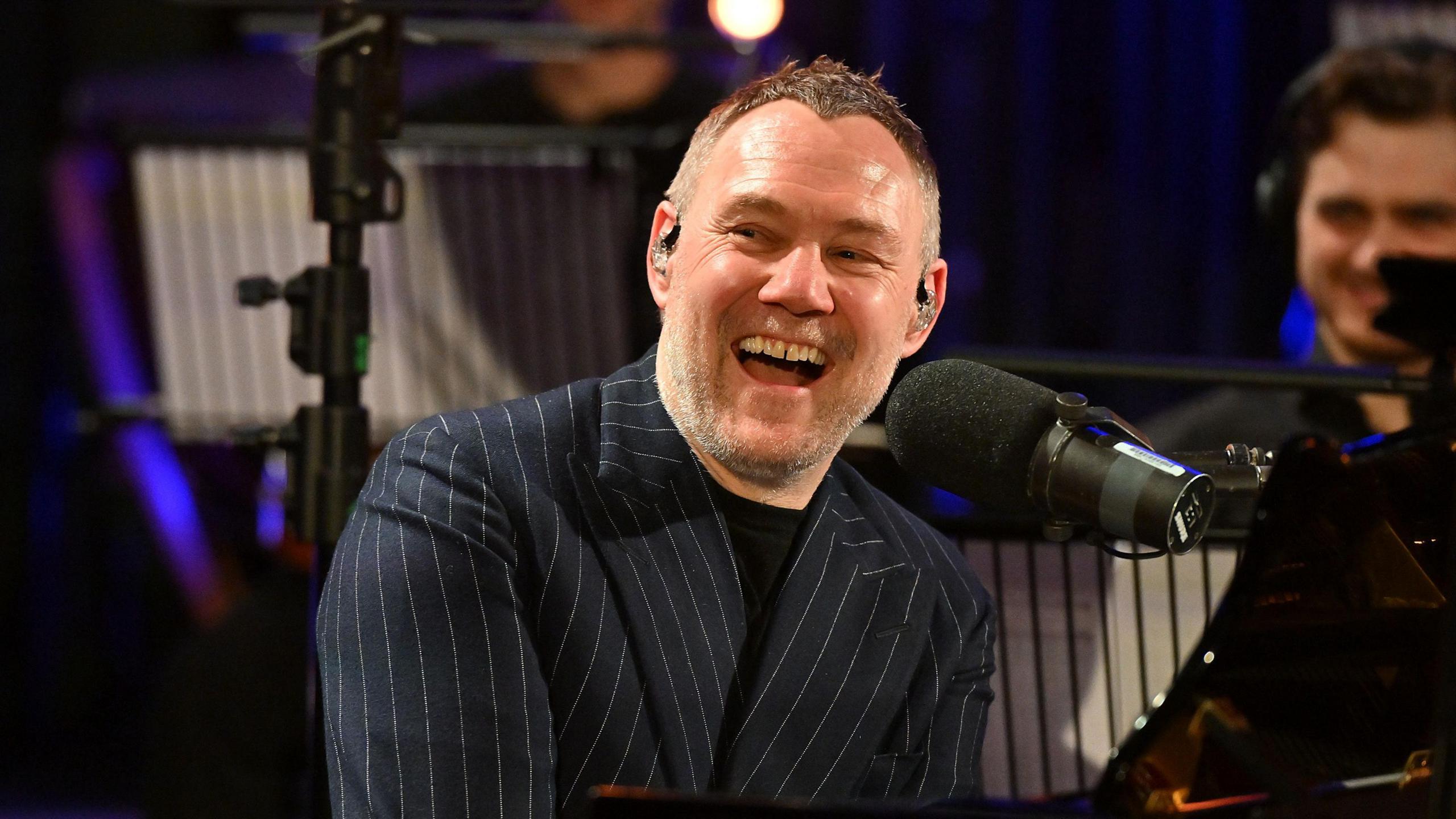 David Gray is sitting at a black piano and laughing into a microphone. He has grey stubble and grey hair. He is wearing a navy blue suit jacket with pin stripes.