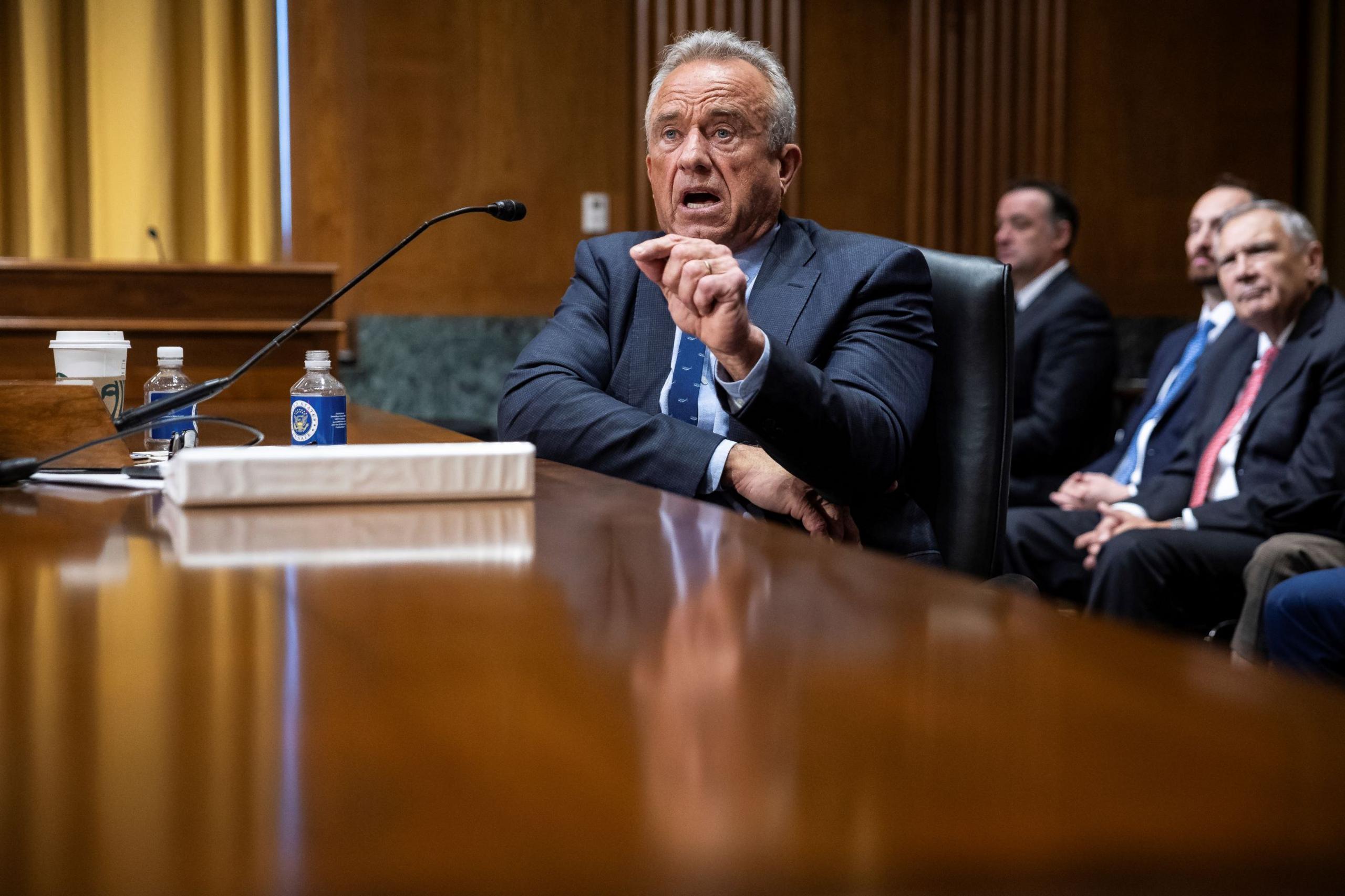 Robert F Kennedy, sits at a wide wooden table. He is wearing a smart, tailored, dark suit. On the table is white folder, some plastic bottles and a white disposable drinks cup. He is speaking animatedly and gesturing with his left hand. In the background, other men in similar dark suits listen.