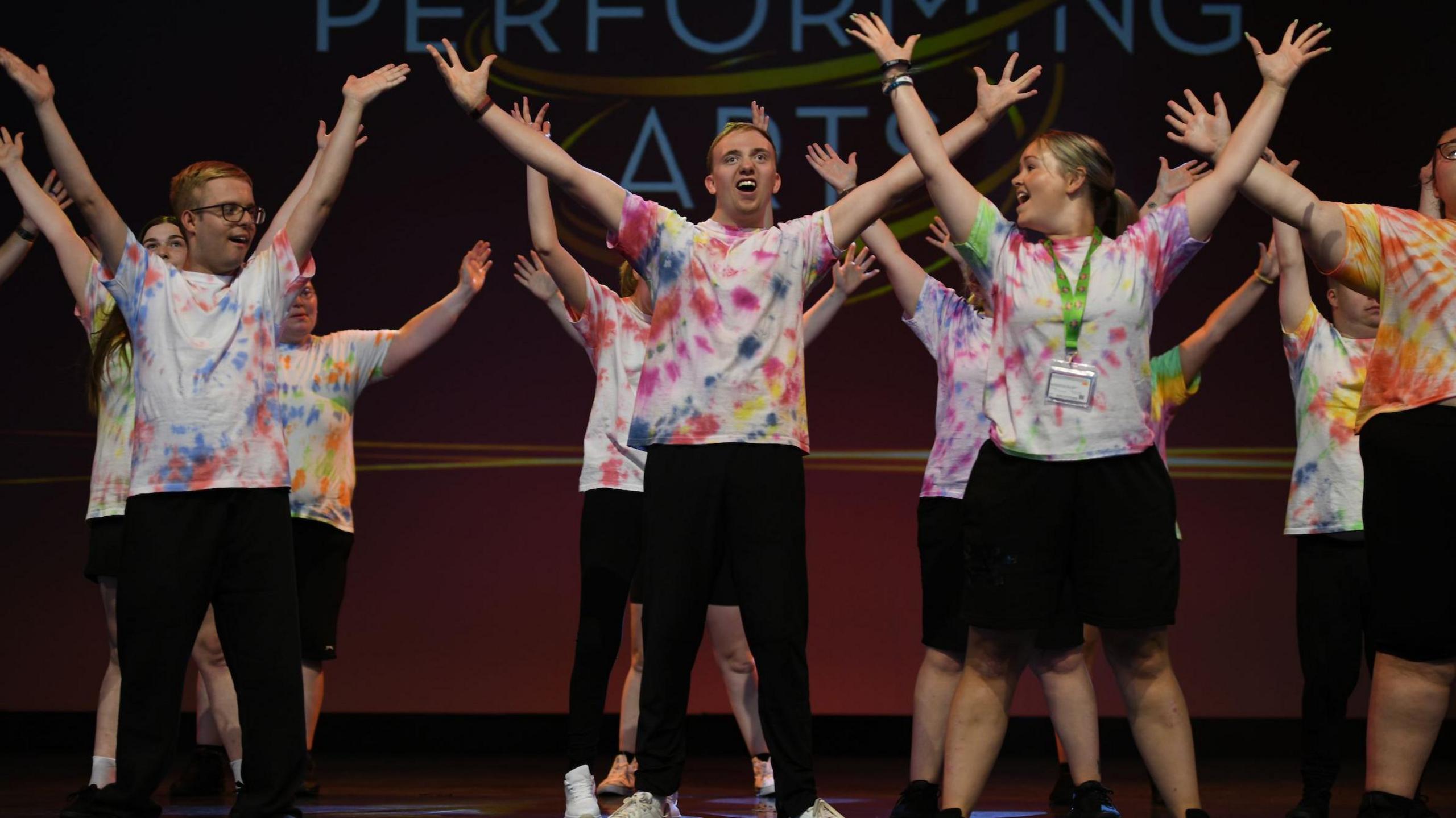 A group of students performing onstage at Disneyland Paris. More than a dozen boys and girls are lifting their legs to music in front of a screen which has "Disney - Performing Arts" on it