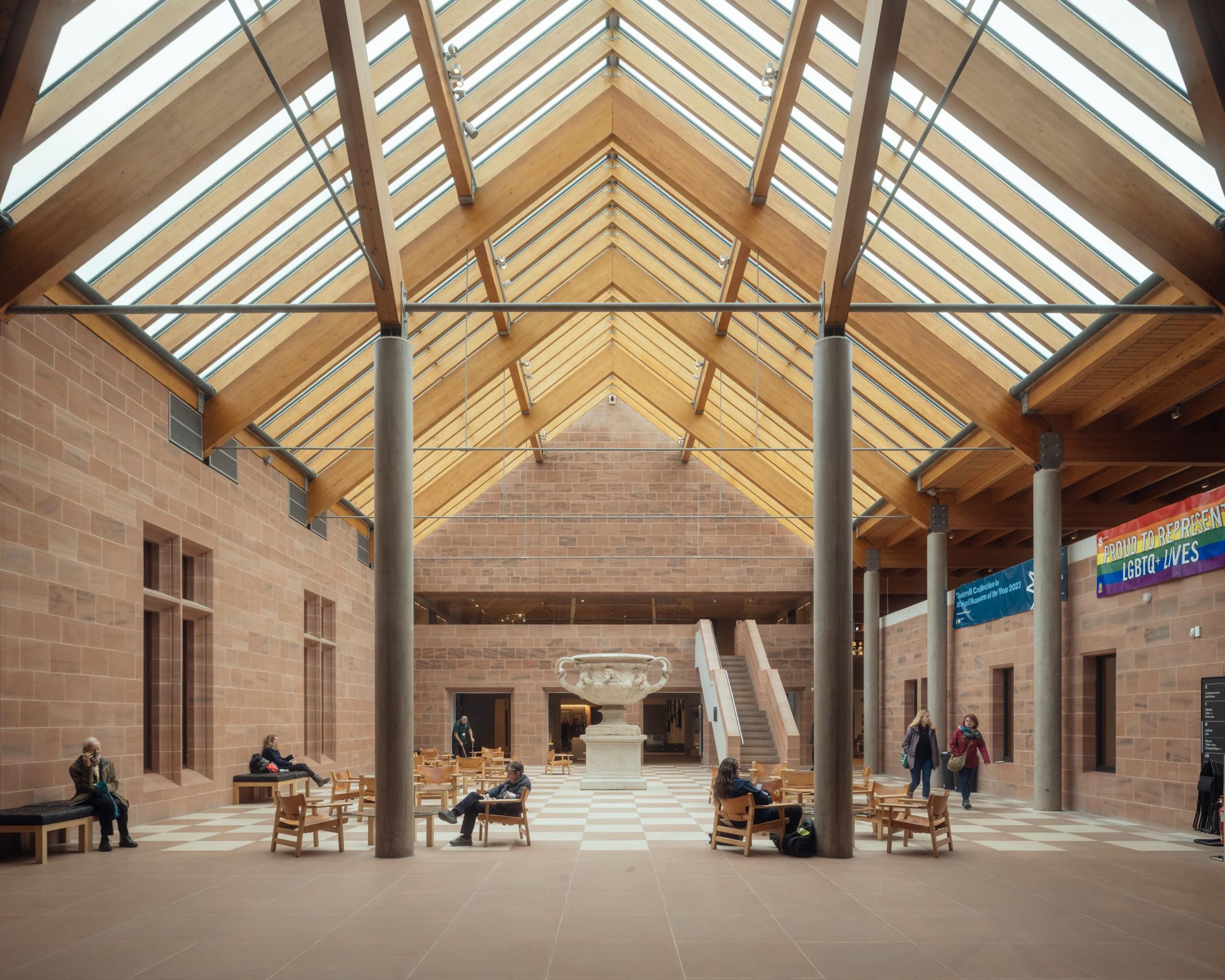 A glass ceiling allows light to flood into a double-height space with chequered floor tiles in beige and white to match the stone walls. People sit at wooden tables and chairs.