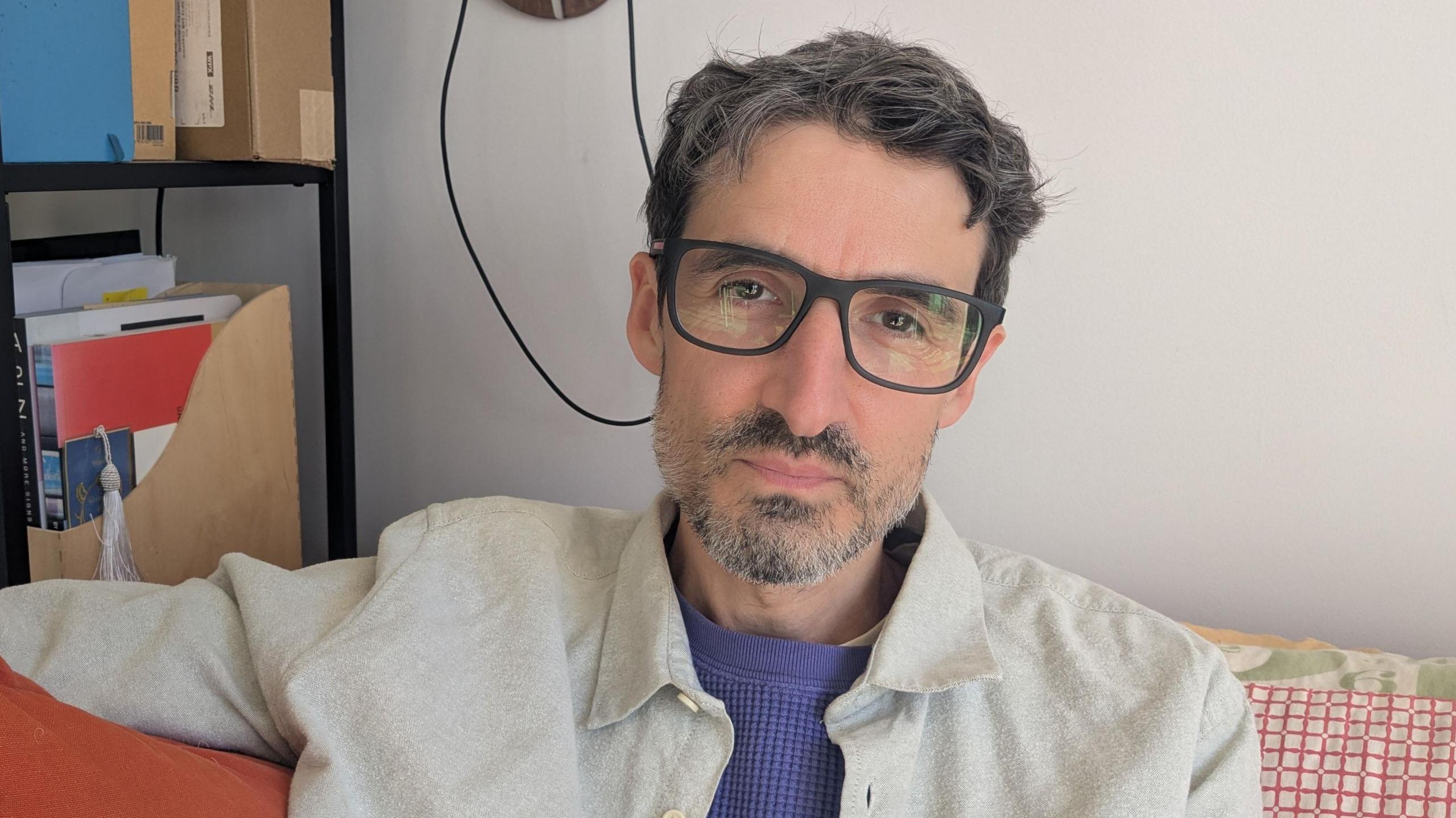 A man in his forties is sitting on a sofa looking at the camera. He's wearing thick-rimmed black glasses and has a short beard. He's wearing a cream shirt over a blue sweater. Next to him is book shelf full of cables and books. 