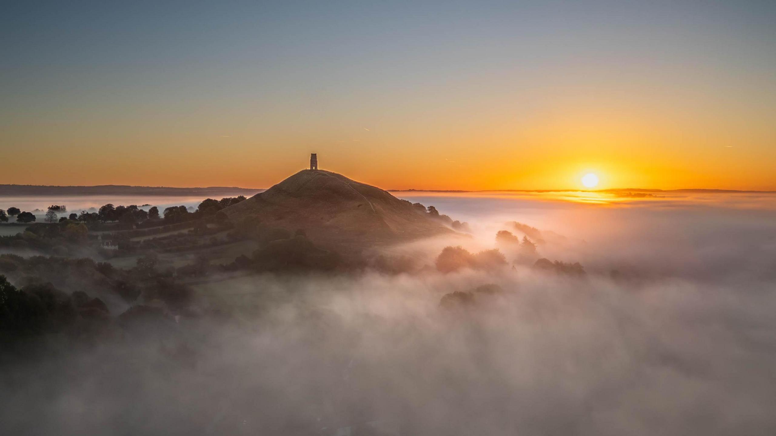 Glastonbury Tor seen from across the valley which is filled up with fog. The sun is rising over the hill.