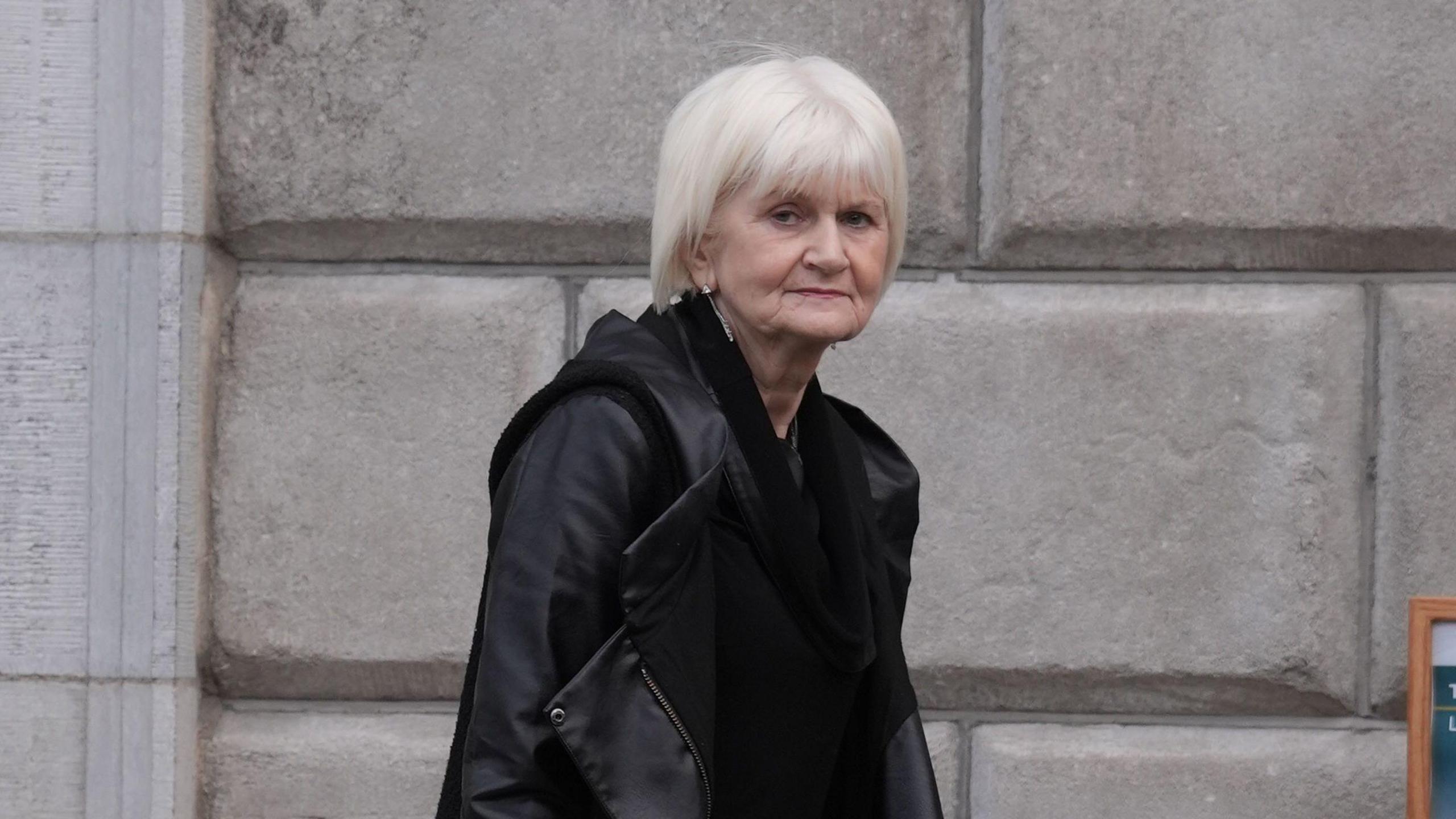 A woman with grey hair, wearing a black coat. She is looking at the camera and standing in front of a grey brick wall. 