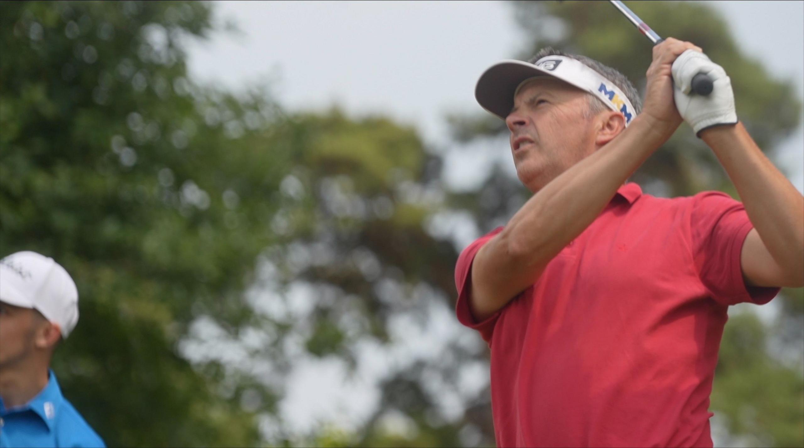 Andrew Marshall wearing a red golf shirt and a white sun visor cap admires his shot on the course at Dereham, with Liam Caffyn in the background watching on.