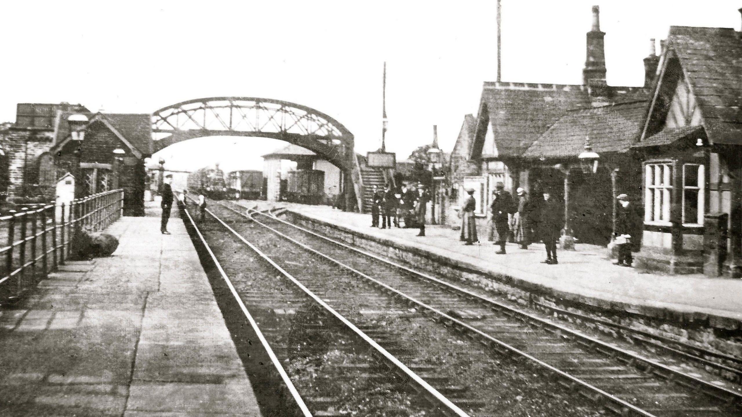 A black and white photograph showing the station platform, with people waiting for the train.