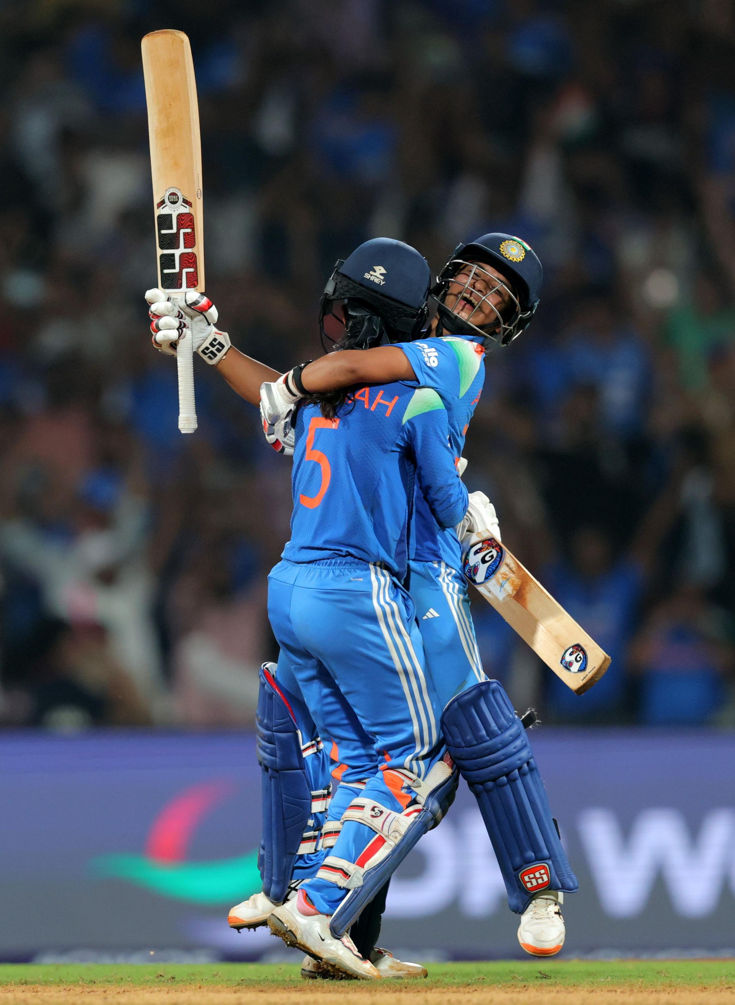 Two cricket players in blue uniforms embrace on the pitch, each holding a bat upright. The scene is set under bright stadium lights with a blurred crowd in the background, suggesting a celebratory moment during a match.