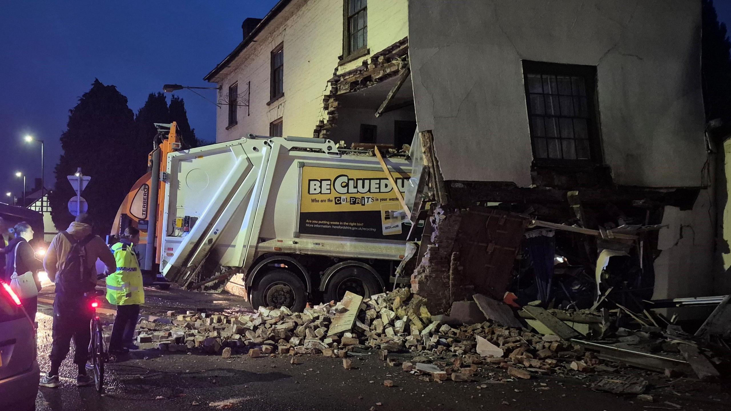 A bin lorry has crashed through the ground floor of a building, with debris lying around the area beside it. There is a silver Ford car in the foreground and a number of people gathered nearby the scene.