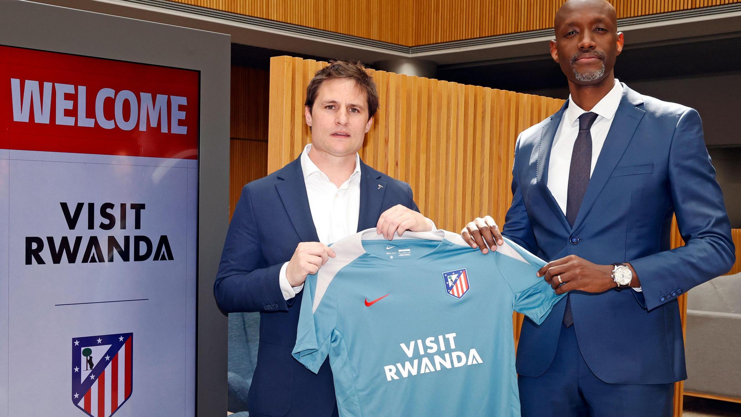 Oscar Mayo of Atletico Madrid and Jean-Guy Afrika of the Rwanda Development Board hold up a light blue Atletico shirt bearing Visit Rwanda on the front