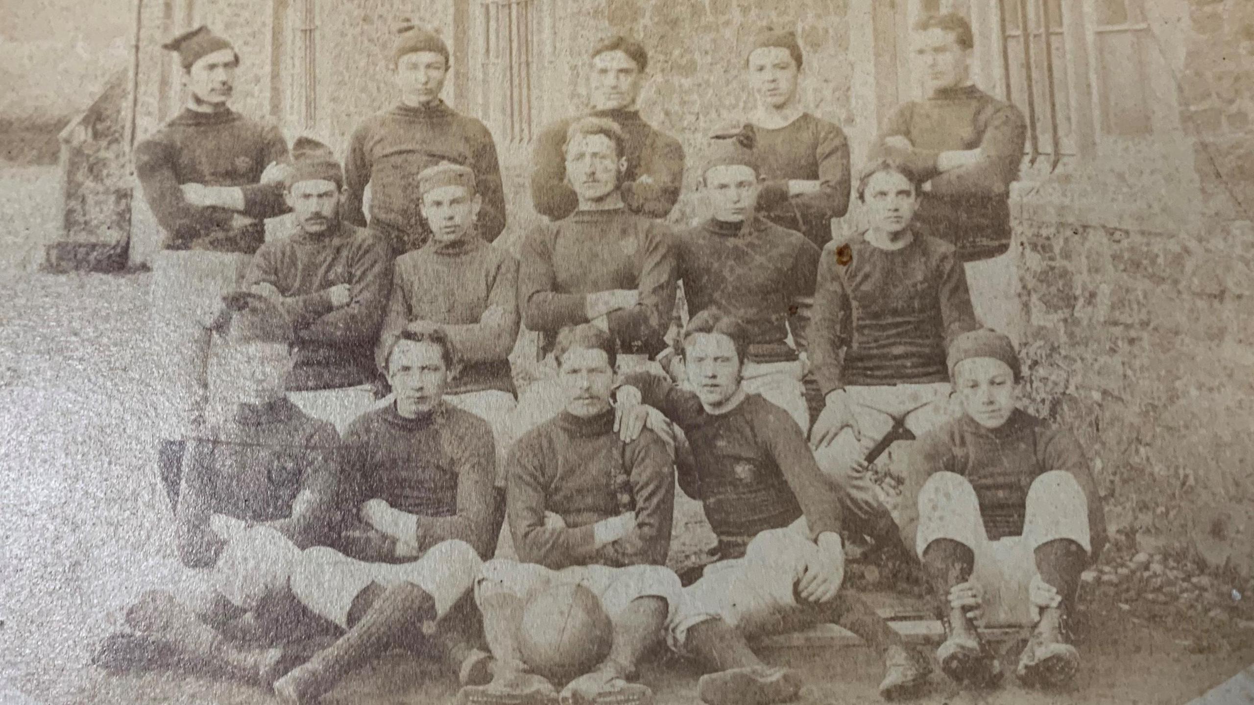 A black and white photo of Llandovery rugby team.
It shows 15 men dressed in long white shorts and long sleeves.
Some of the men are wearing little floppy hats.