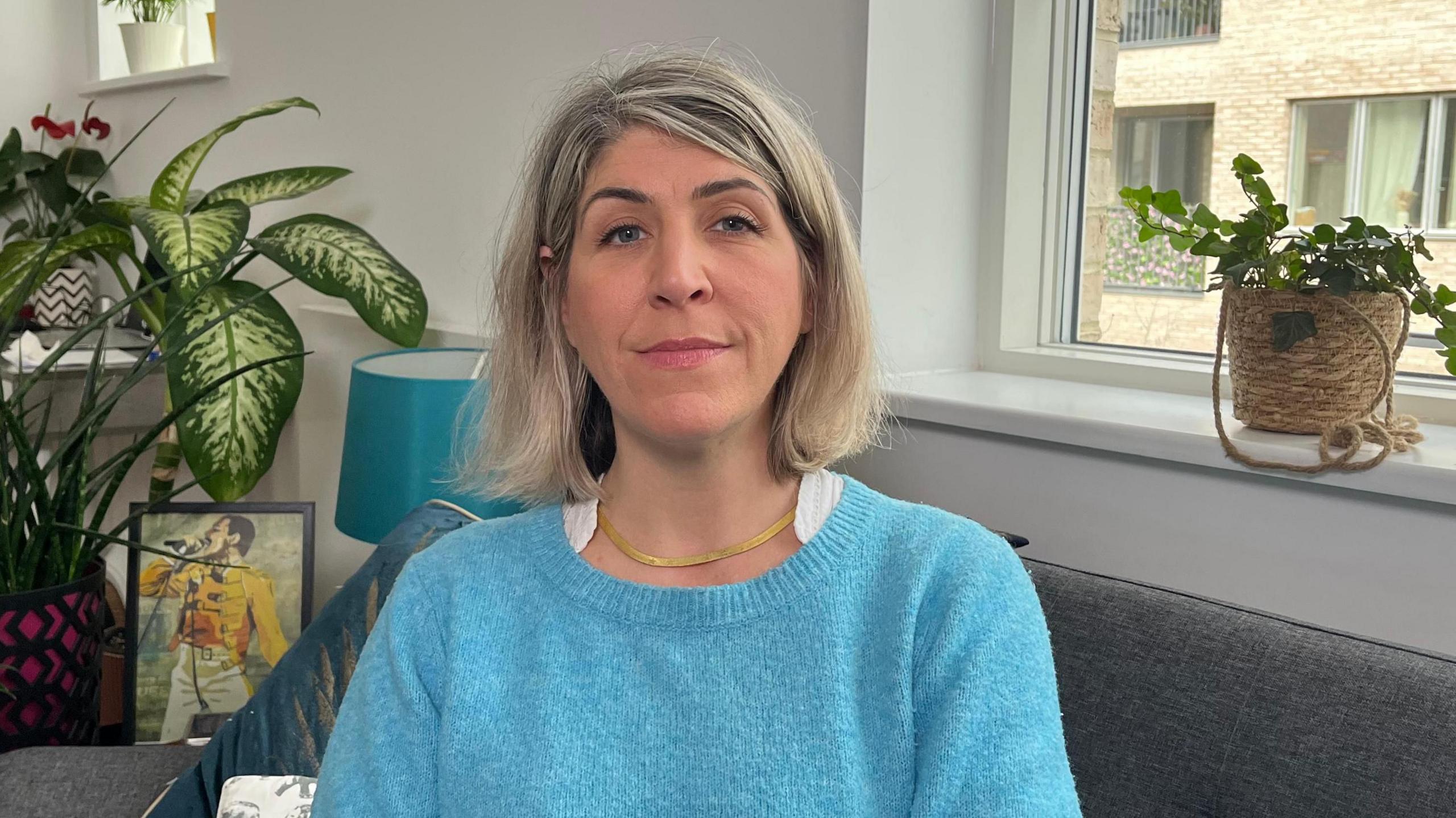 A woman with shoulder-length light hair wearing a blue sweater sits on a grey sofa with her arms crossed